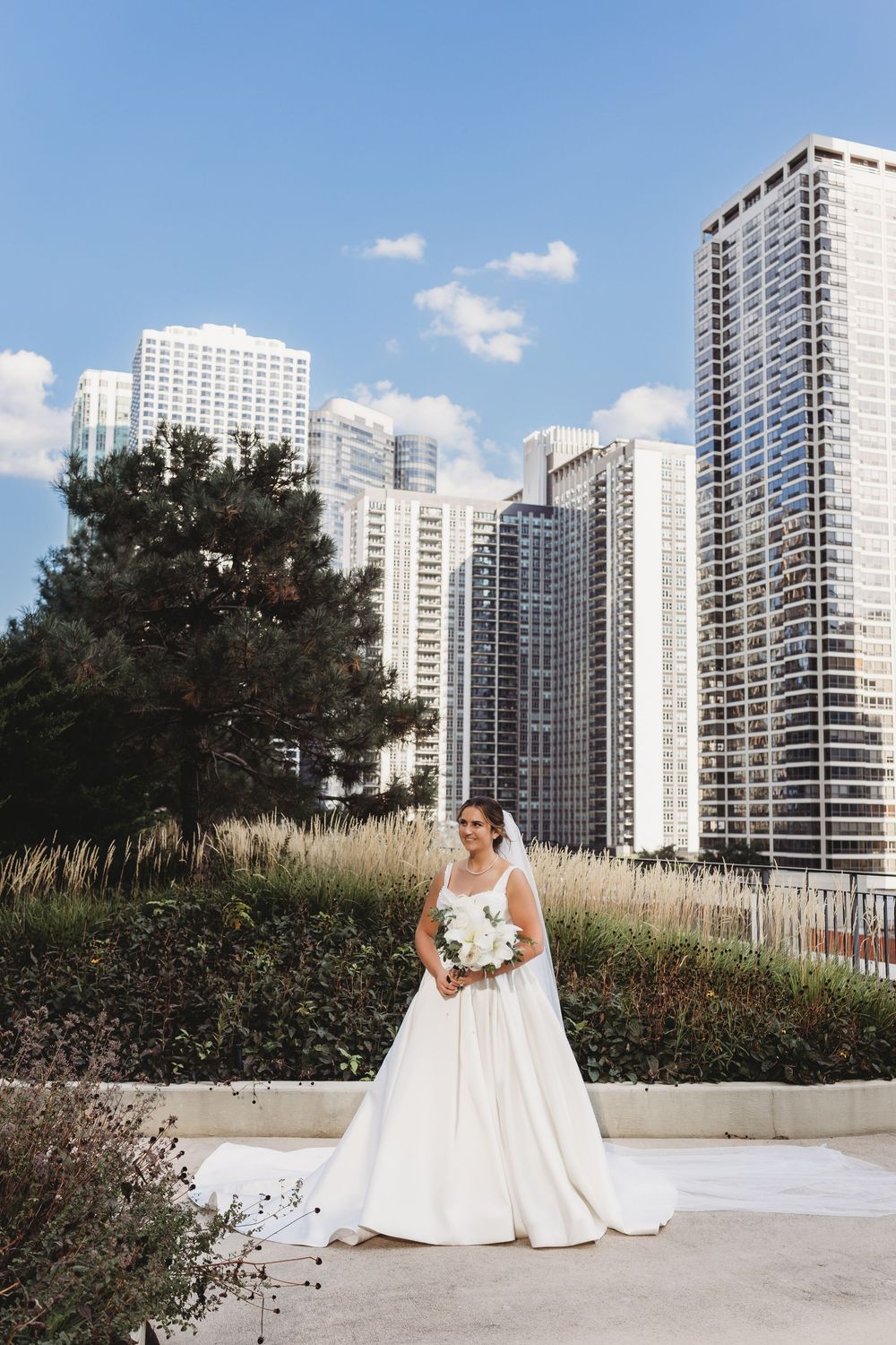 A bride in a flowing white wedding gown poses against a backdrop of modern high-rise buildings and tall grasses.