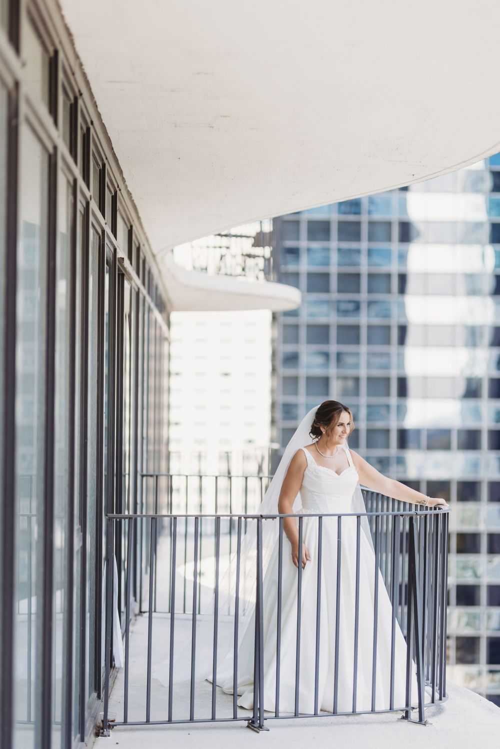 A bride in a white wedding dress poses gracefully along a modern building's glass-walled balcony.