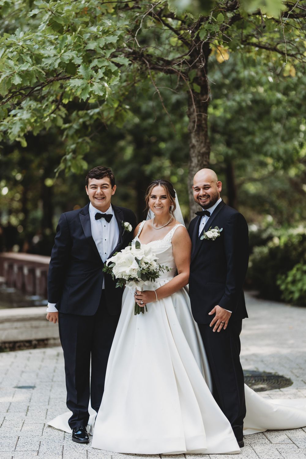 A wedding party poses outdoors on a stone path under green trees with the bride holding a white bouquet.