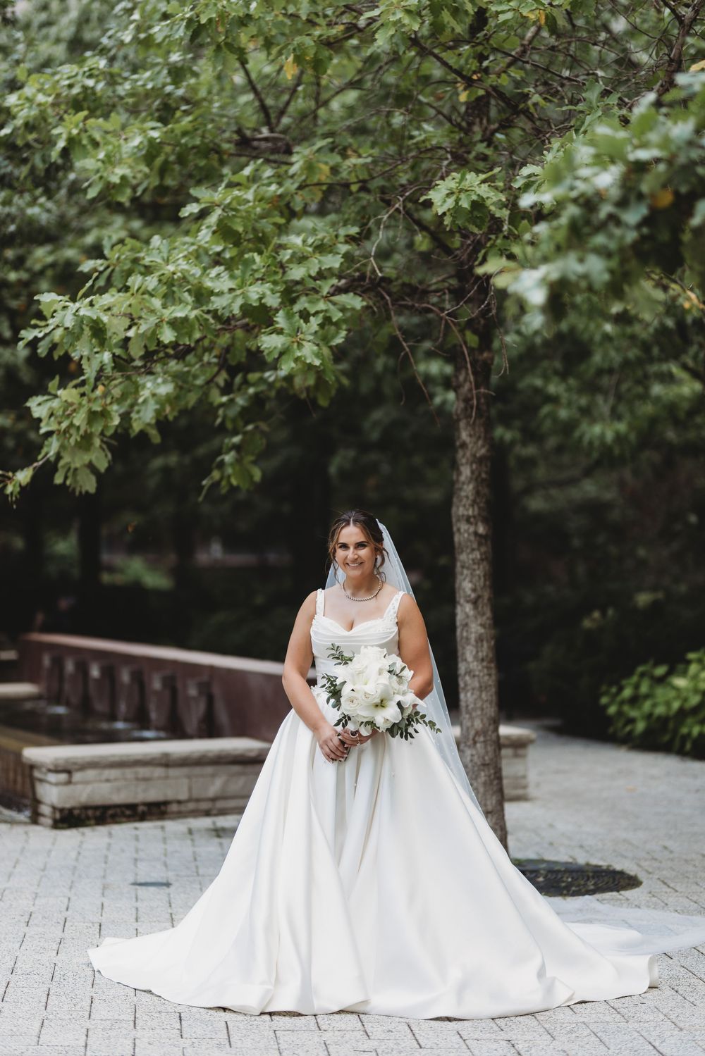 A bride in a flowing white wedding gown poses gracefully in a garden setting with trees and stone pathways.
