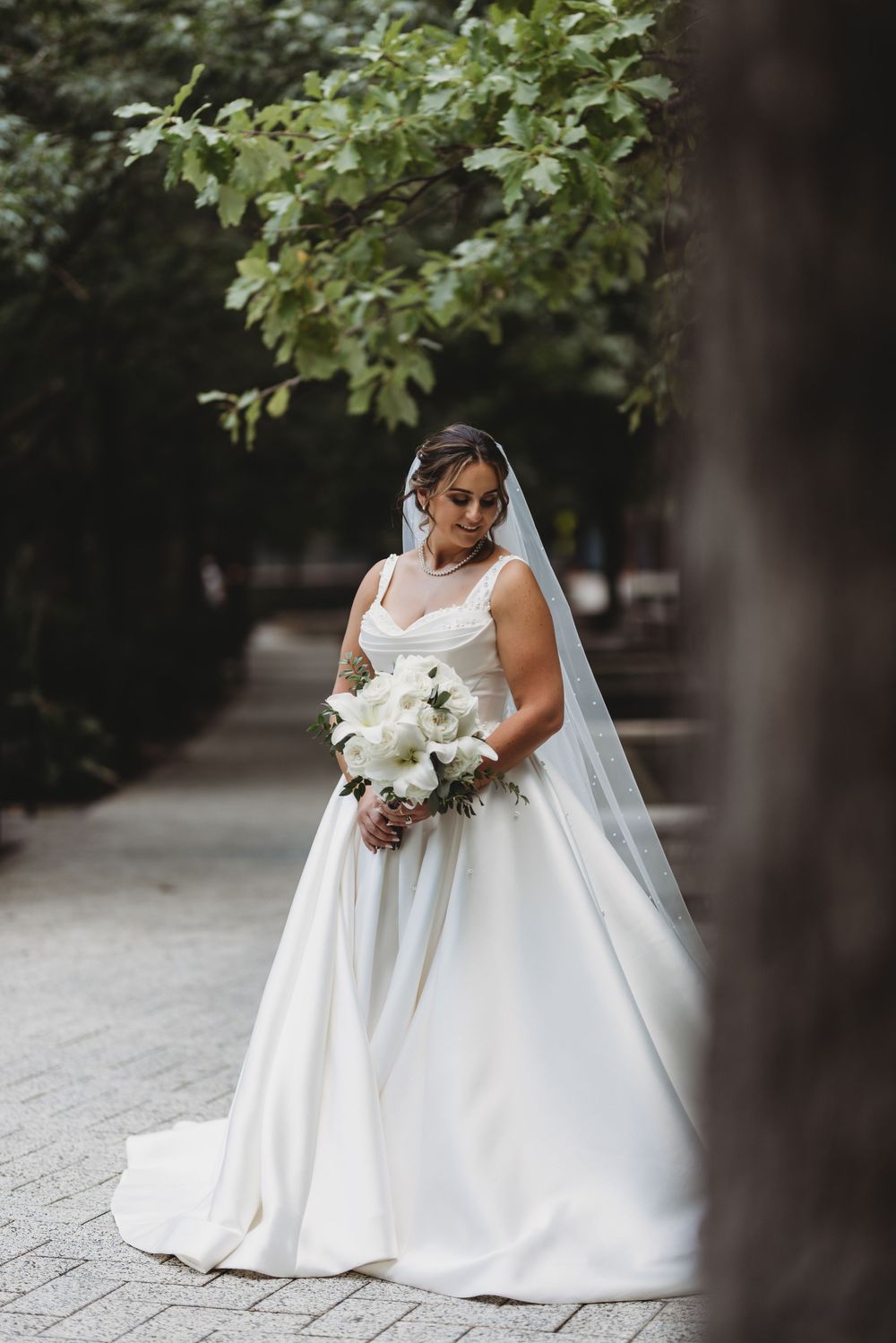 A bride in an elegant white satin ballgown holds a bouquet of white flowers in an outdoor garden setting.
