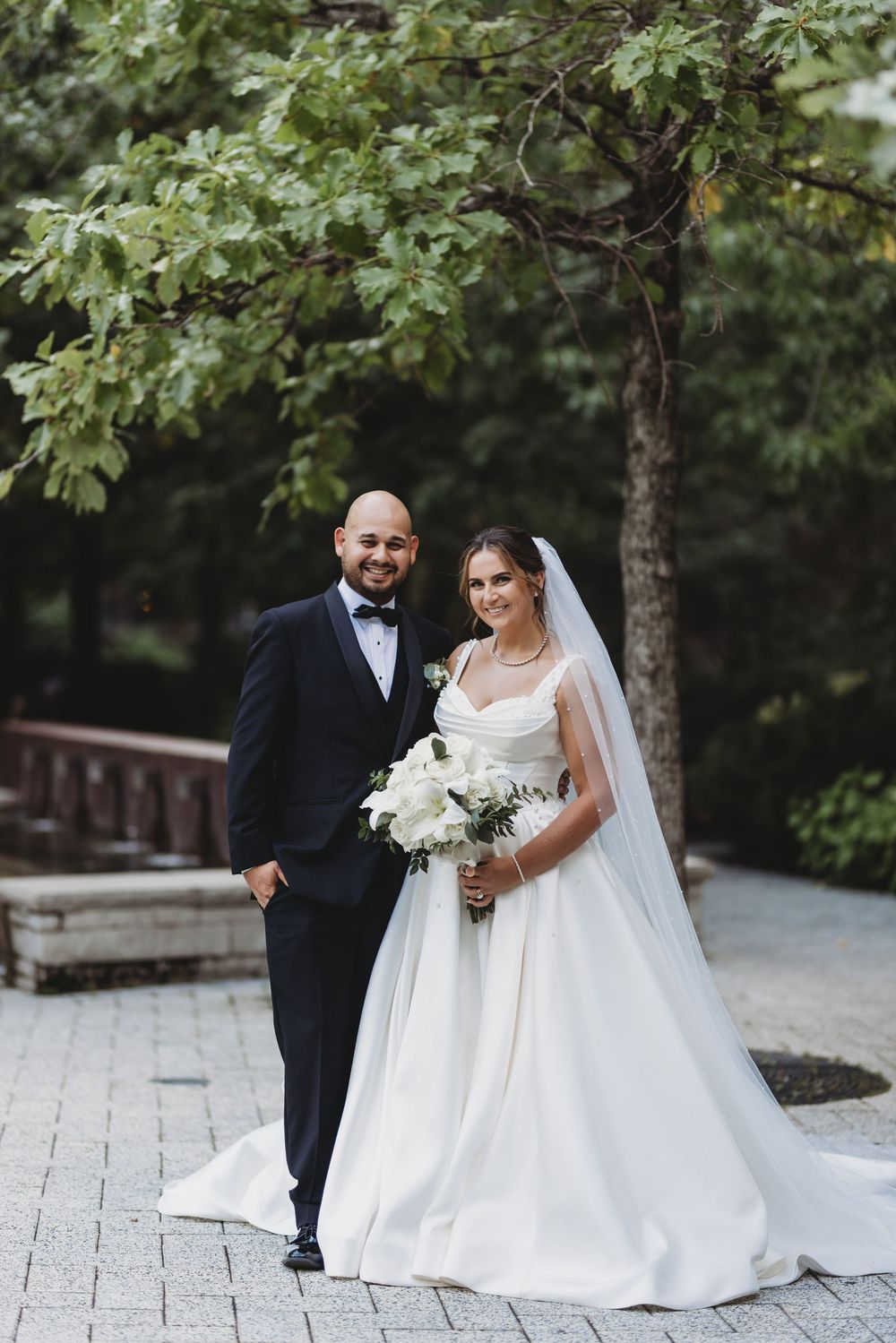 A couple in formal wedding attire shares joyful moments on a stone pathway surrounded by lush green trees.