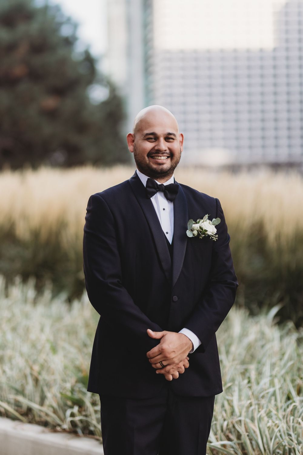 A groom in a classic black tuxedo poses outdoors with city architecture in the background.