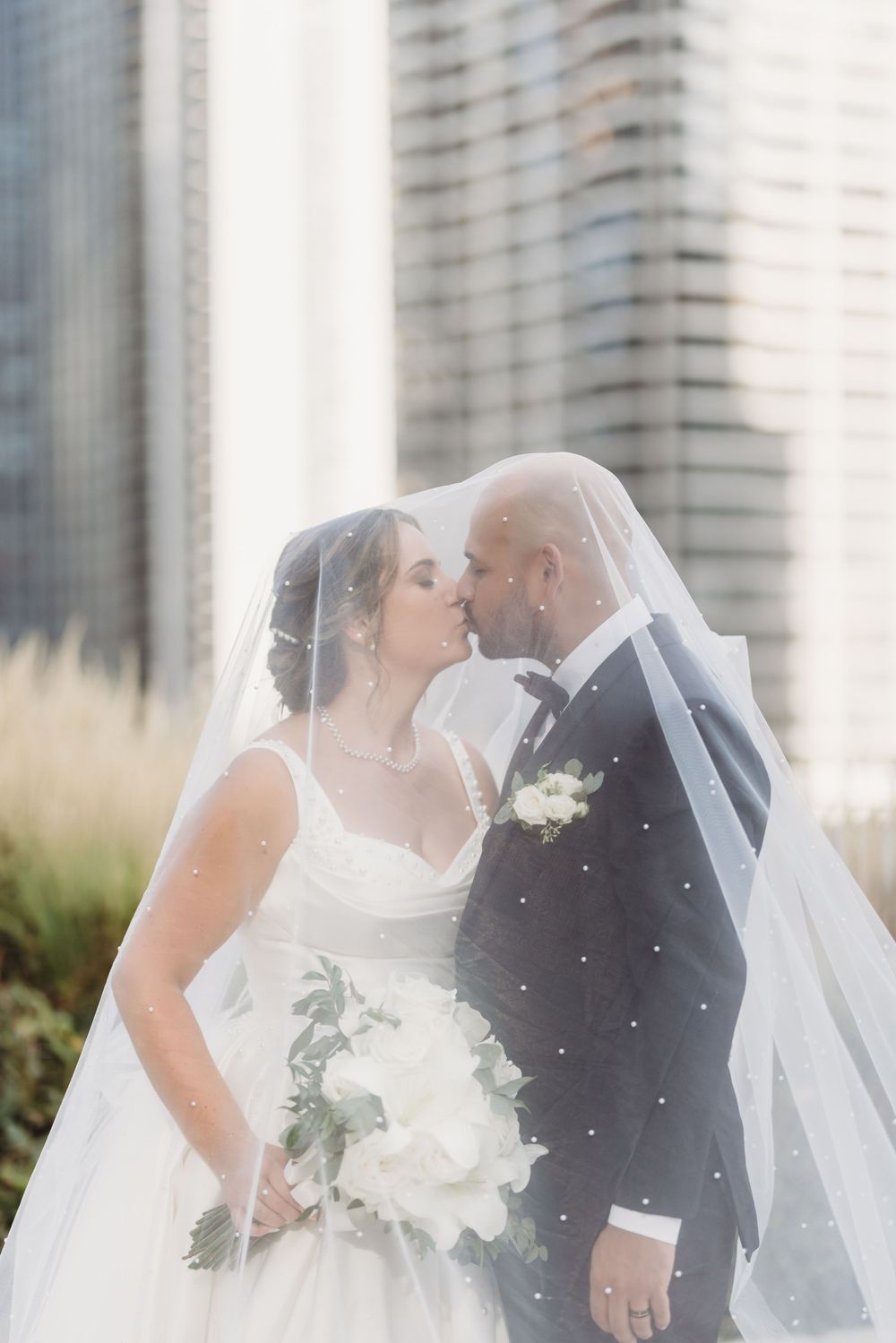A romantic wedding couple shares an intimate moment under a flowing white veil against an urban backdrop.