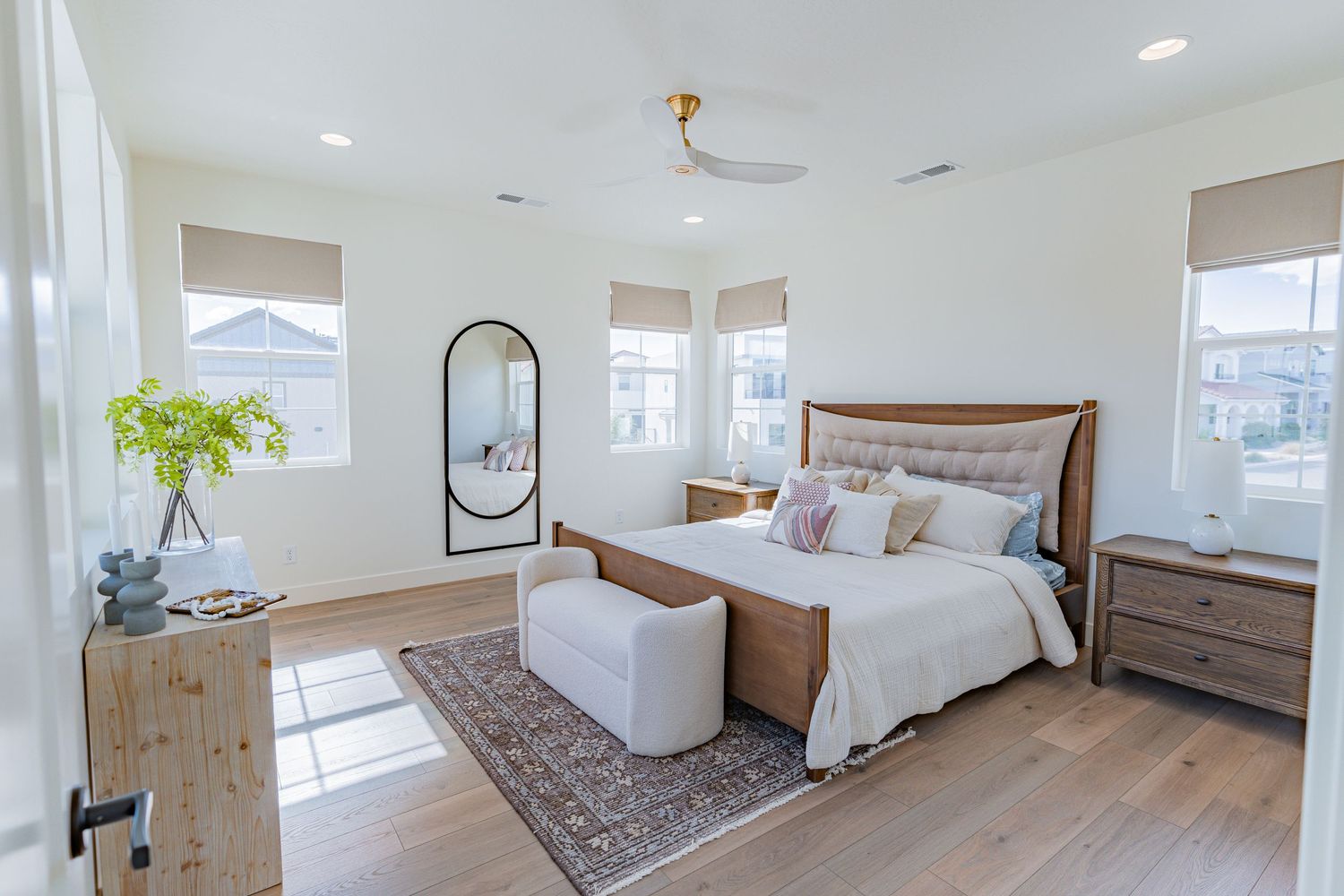 Bright modern bedroom with wood-frame bed, white armchair, hardwood floors, and large windows in a neutral palette.
