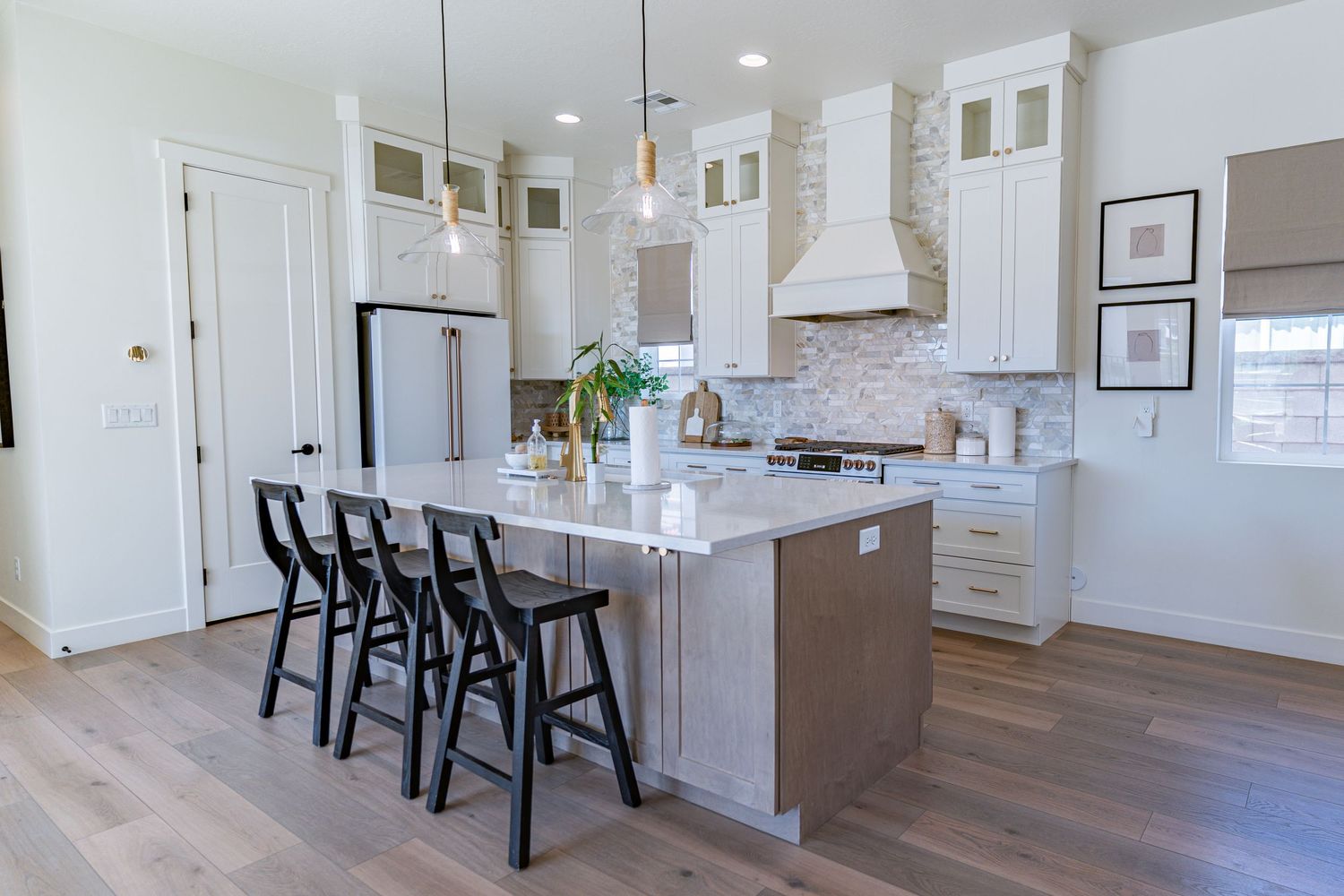 Modern white kitchen with large island, black bar stools, stainless steel appliances, and hardwood floors.