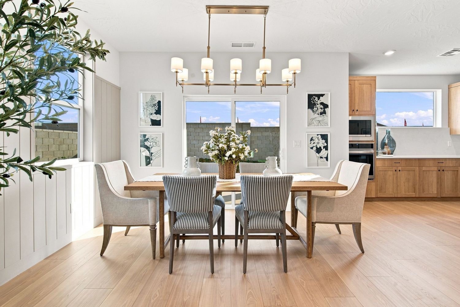 Bright dining room with a rectangular wood table, upholstered chairs, gold chandelier, and large windows on light hardwood floors.