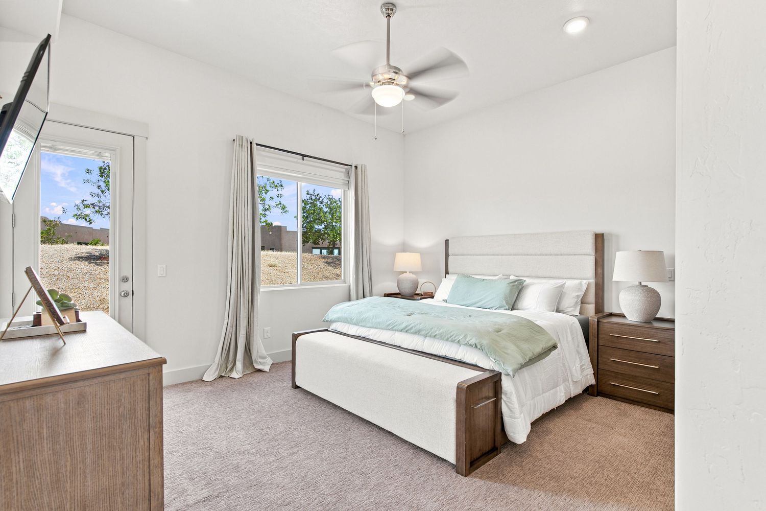 Bright master bedroom with ceiling fan, plush carpet, white upholstered bed, wood nightstands, and natural light from large windows.