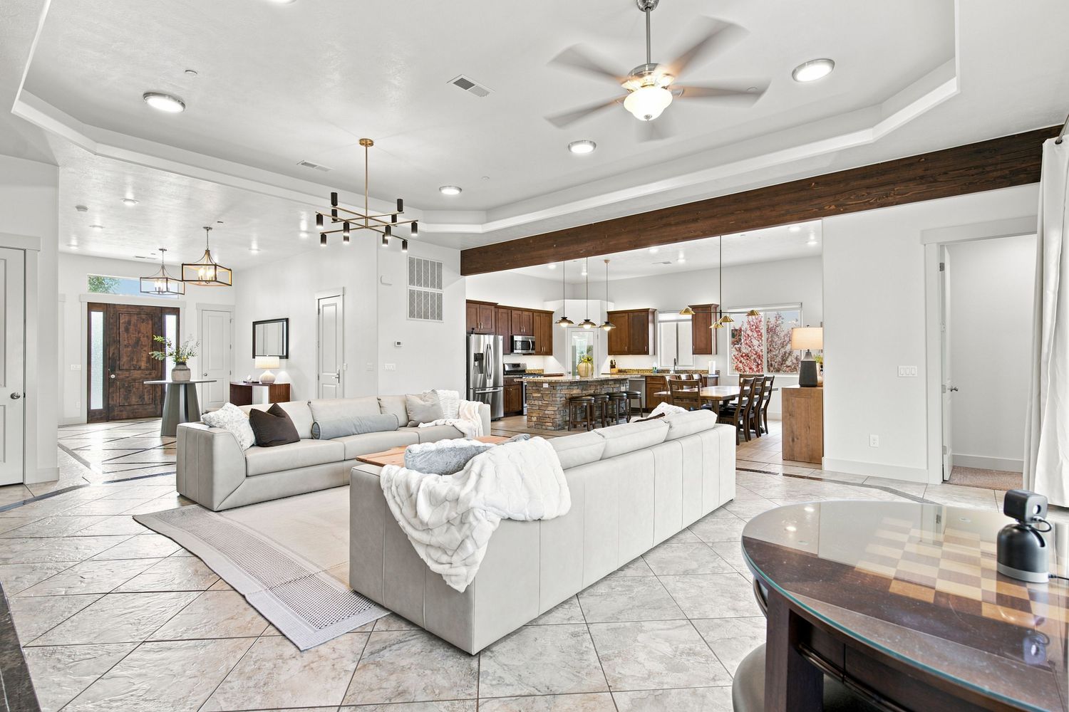Spacious open-concept living room with tray ceiling, ceiling fan, tile floors, and view into modern kitchen.