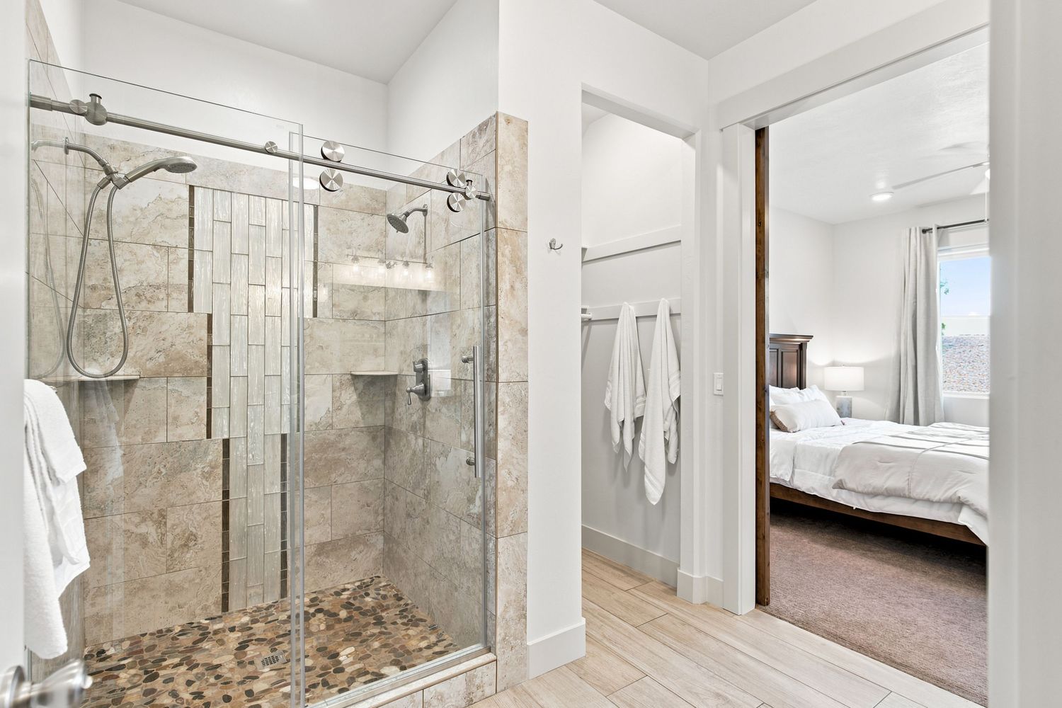 Modern ensuite bathroom featuring a walk-in tile shower with glass doors, white towels, and view into bright bedroom.