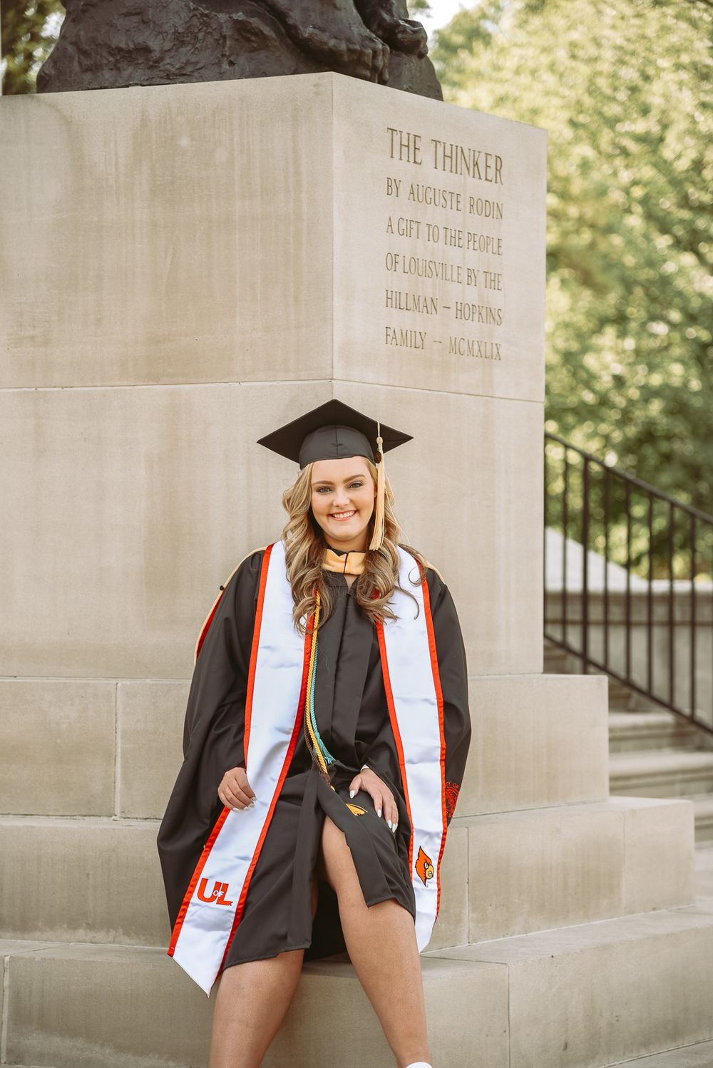 Graduate poses near stone wall with 'The Thinker' inscription in academic regalia.