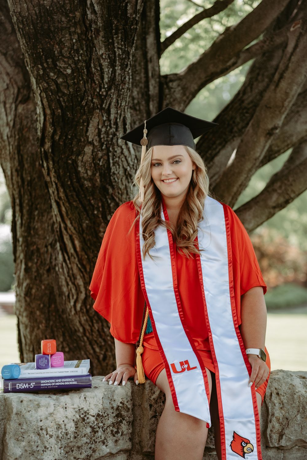 Graduate in red stole and cap poses against tree trunk with textbooks.