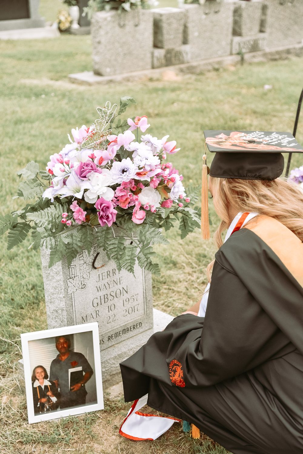 Graduate in black gown kneels by decorated gravestone with pink flowers.