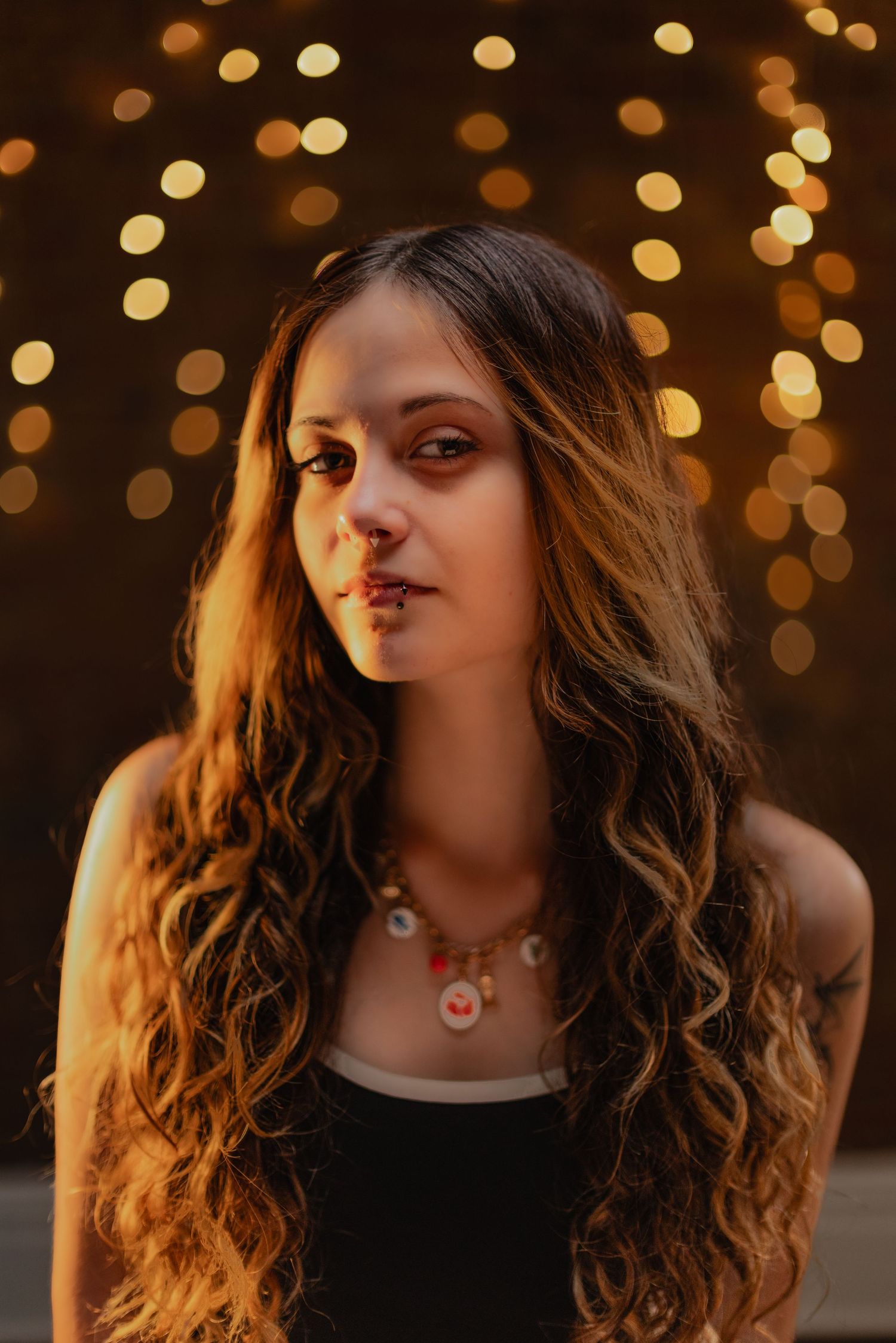 Portrait with long wavy brown hair against warm bokeh lights wearing black top and necklace in golden lighting.
