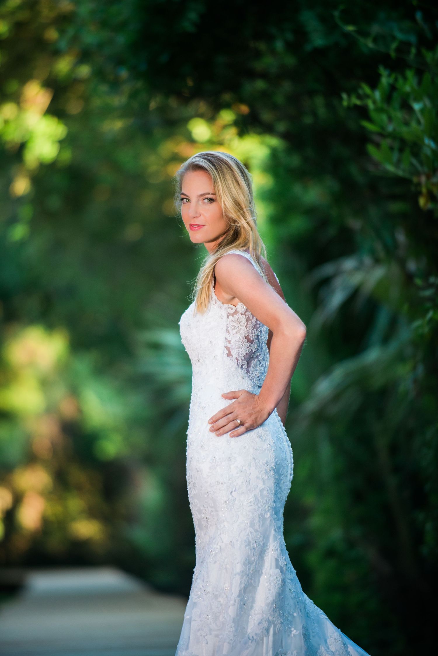 A woman in a white sequined wedding dress poses against a lush green outdoor backdrop.