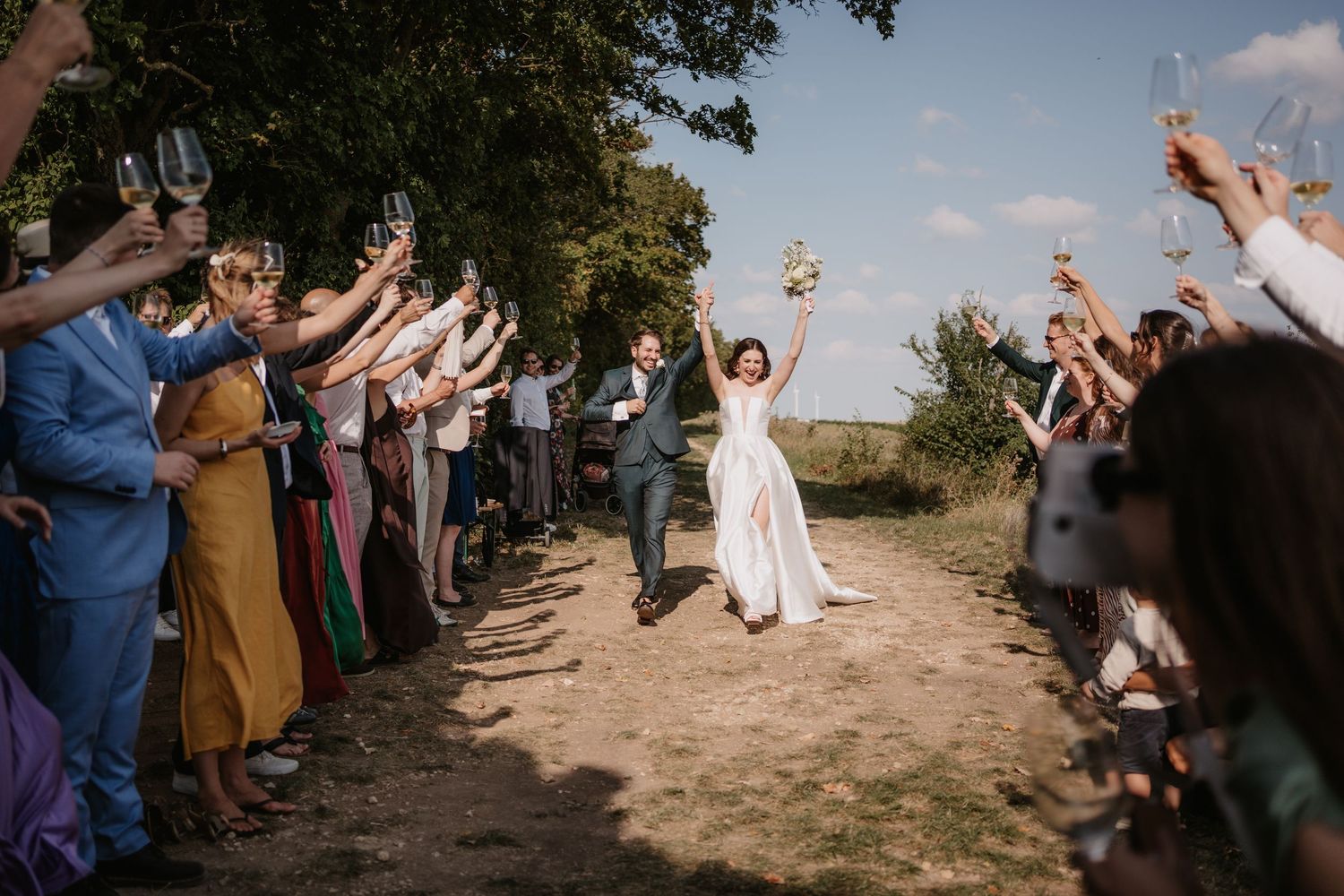 Joyful wedding party celebrates outdoors with raised arms and big smiles on a sunny day.
