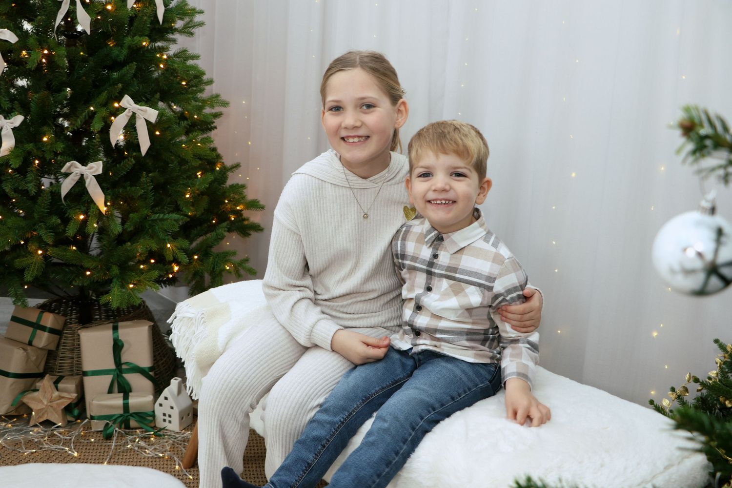 Children in cozy holiday pajamas relaxing on white bedding with Christmas decor and twinkling lights in background.