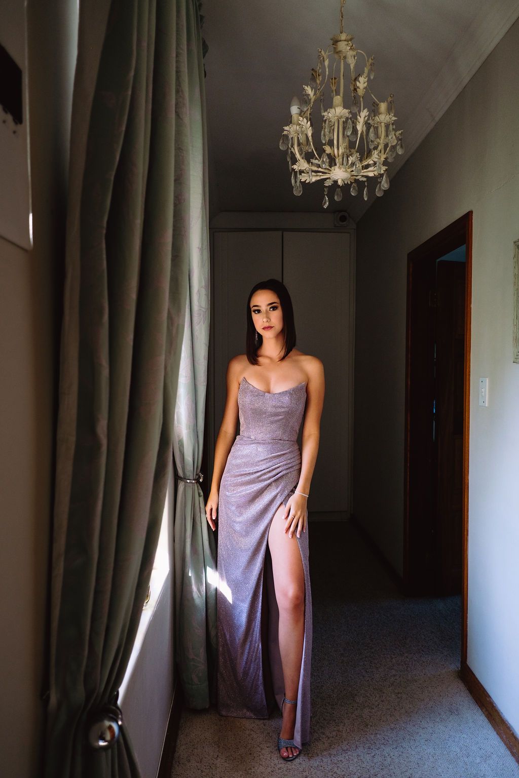 Matric farewell portrait of a young woman in a purple dress standing by a window under a chandelier, photographed in Kwambonambi