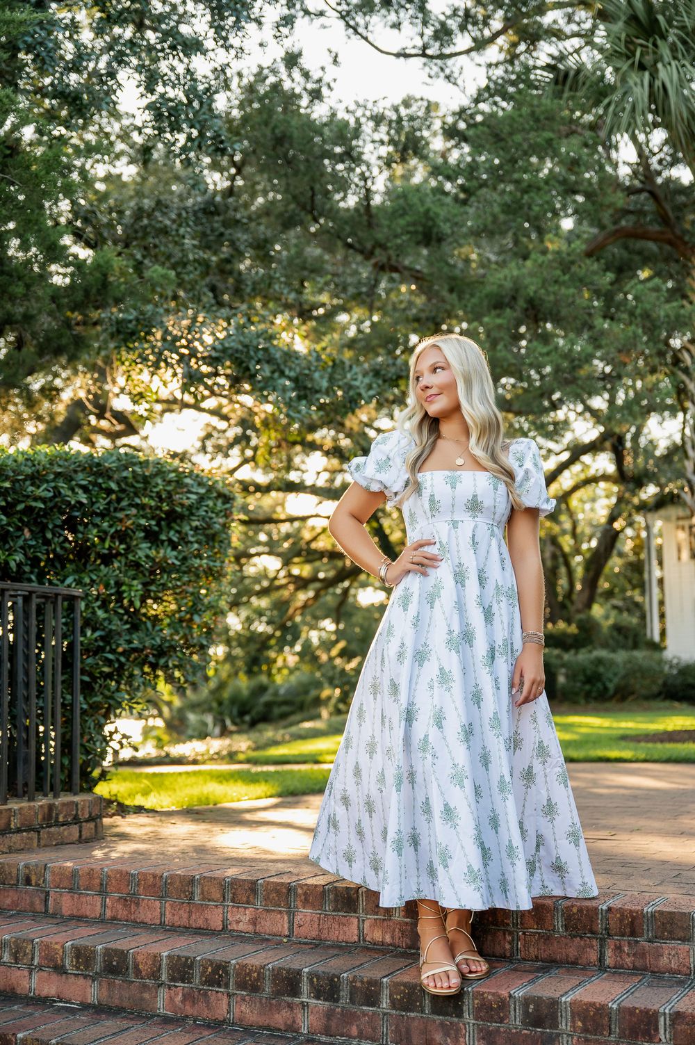 Woman in white patterned dress poses on brick steps surrounded by greenery.