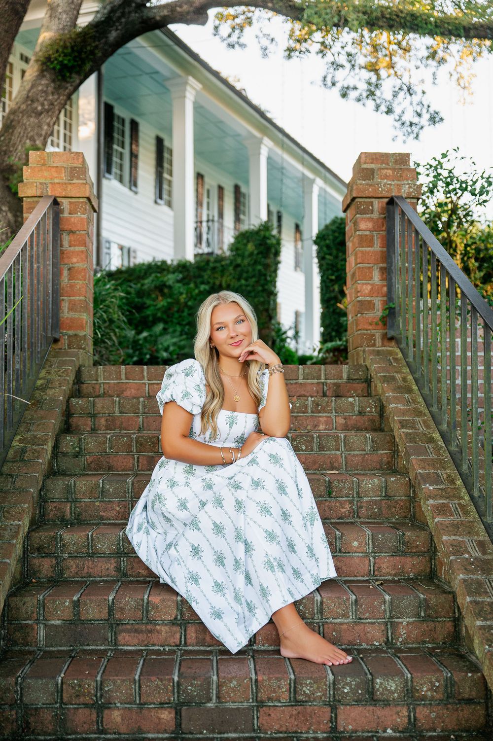 Woman in floral dress poses on brick steps with white colonial architecture backdrop.