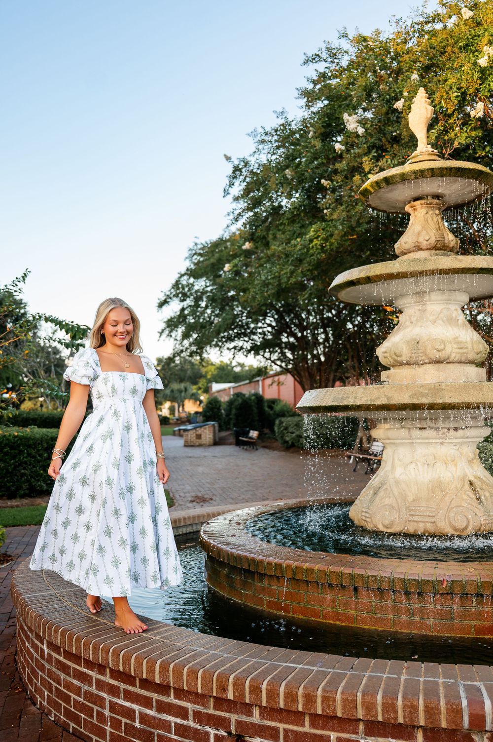 White sundress flowing near a classic brick fountain in a garden setting at sunset.