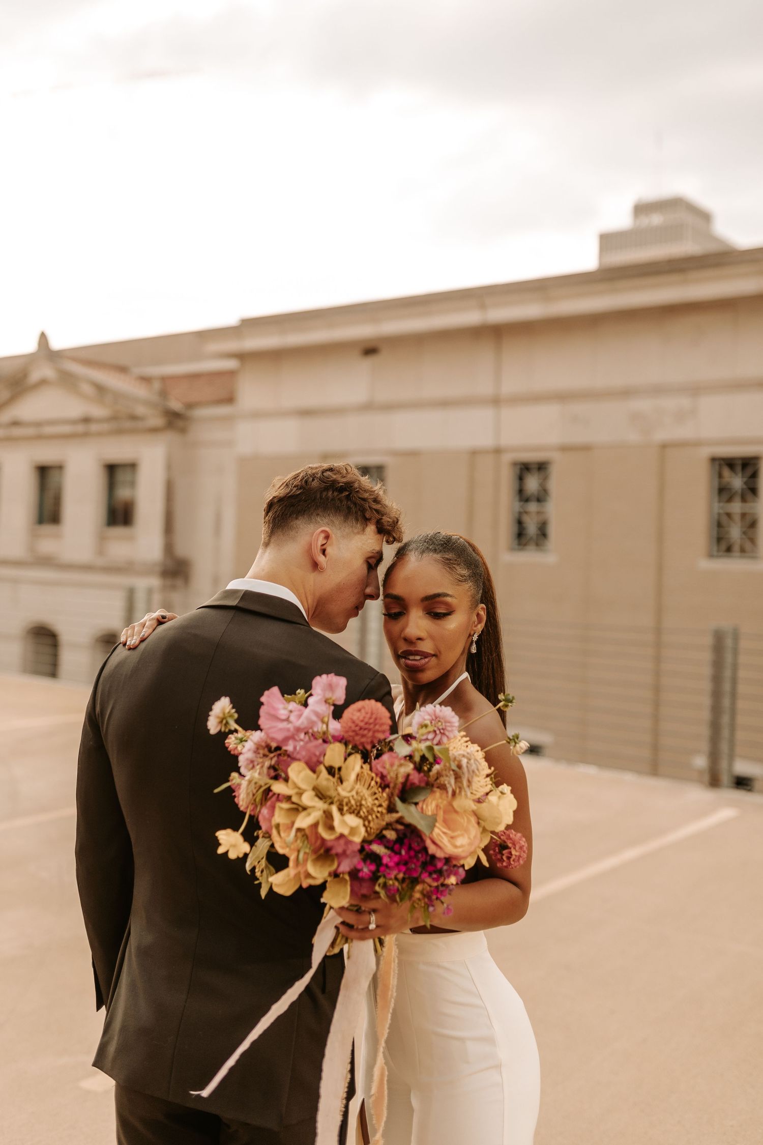 A romantic couple shares an intimate moment on a rooftop with a city skyline backdrop at sunset.