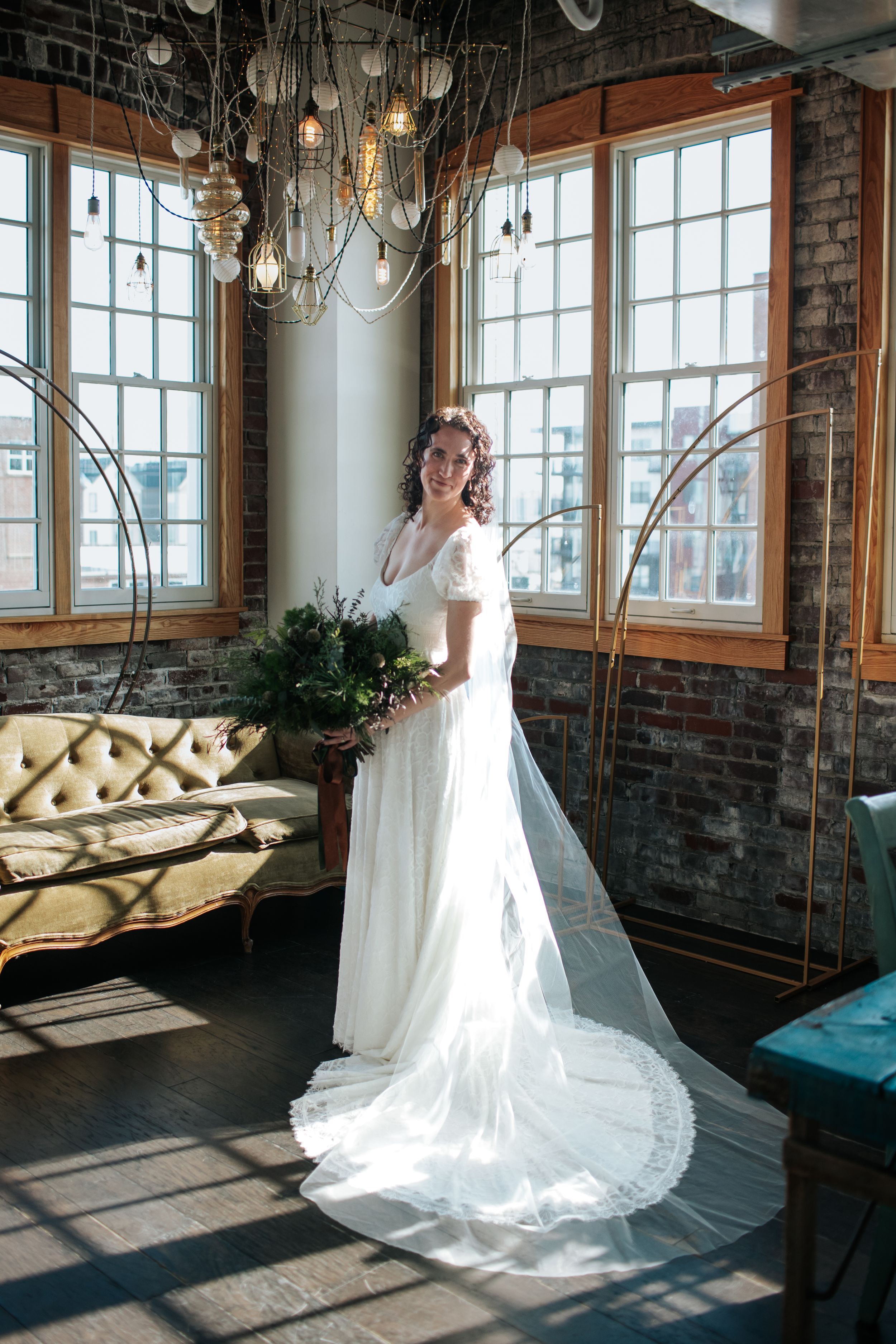A white wedding dress with a flowing train is showcased in a rustic industrial venue with exposed brick and large windows.