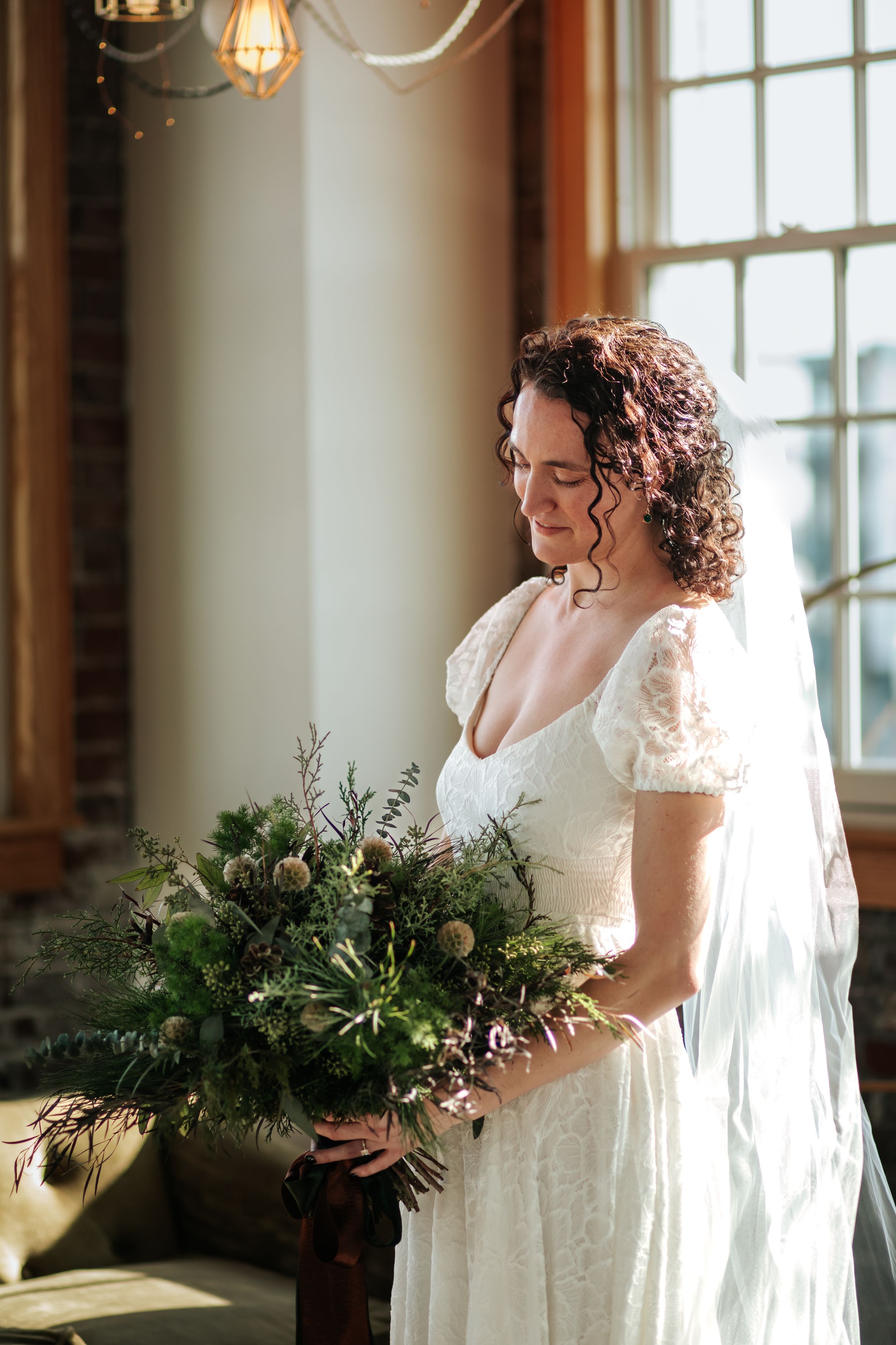 Two brides in elegant white wedding dresses share a tender moment near sunlit windows with lush greenery bouquets.
