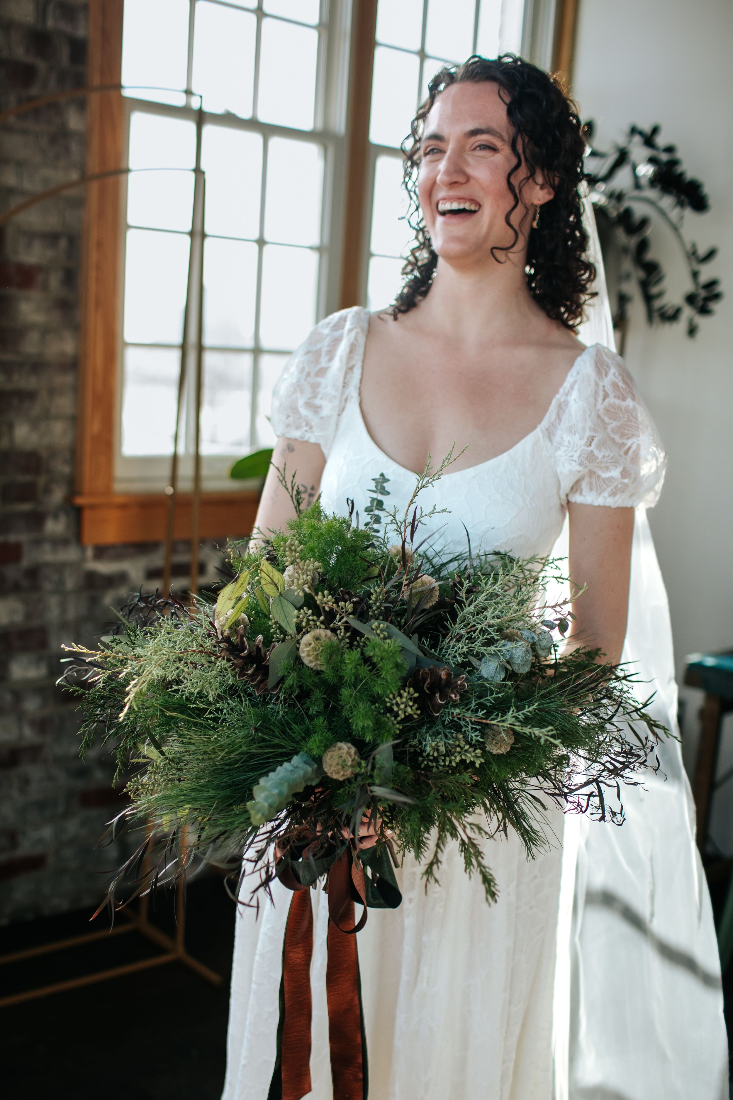 A person in a white wedding dress holds a lush evergreen and floral bouquet near a bright window.