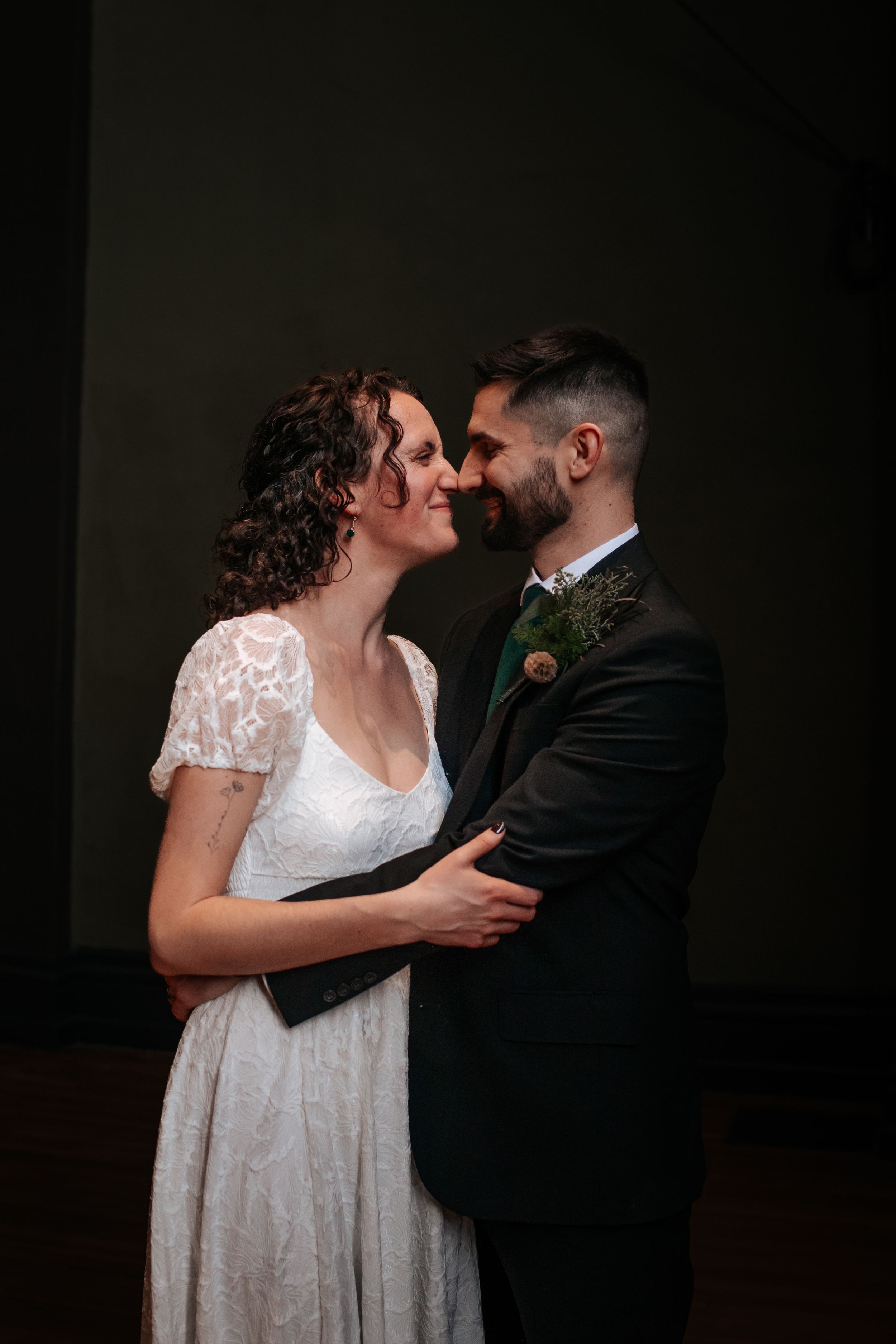 A romantic wedding couple in formal attire share an intimate moment in a dark, moody portrait series.