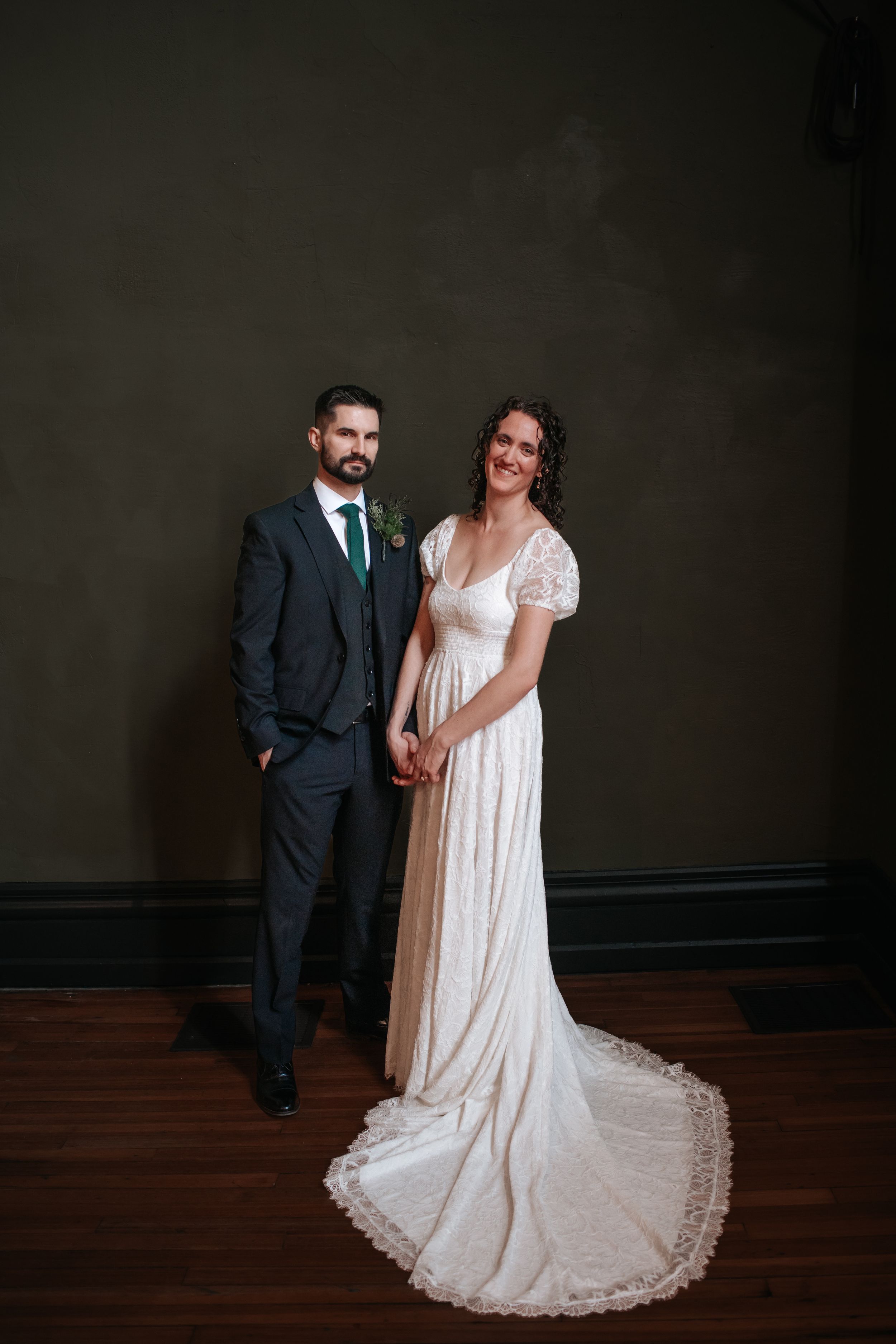 A couple poses for a formal portrait against a dark wall, wearing a dark suit and flowing white wedding gown.