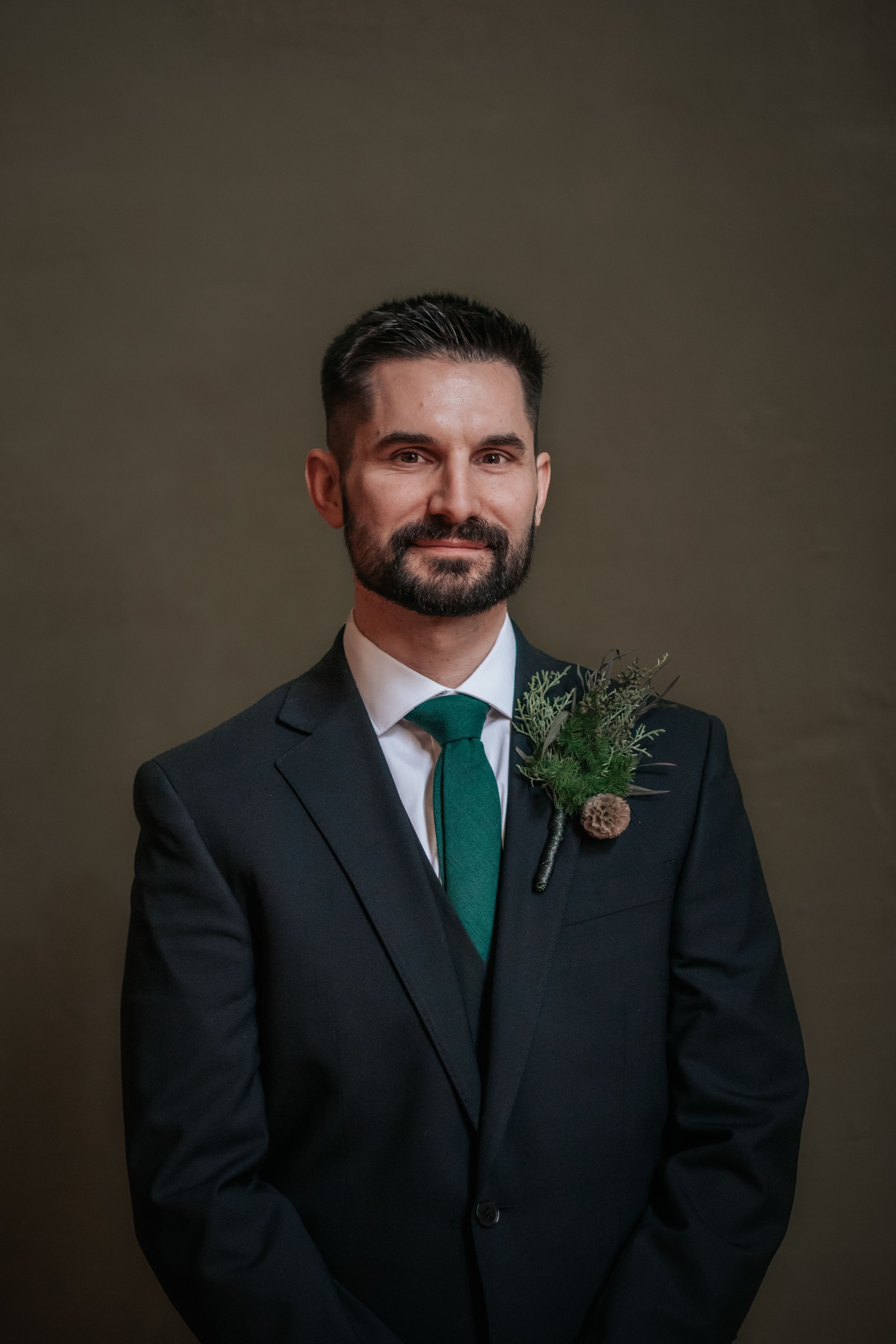 A groom in a dark suit and emerald green tie smiles while wearing a boutonniere against a dark background.