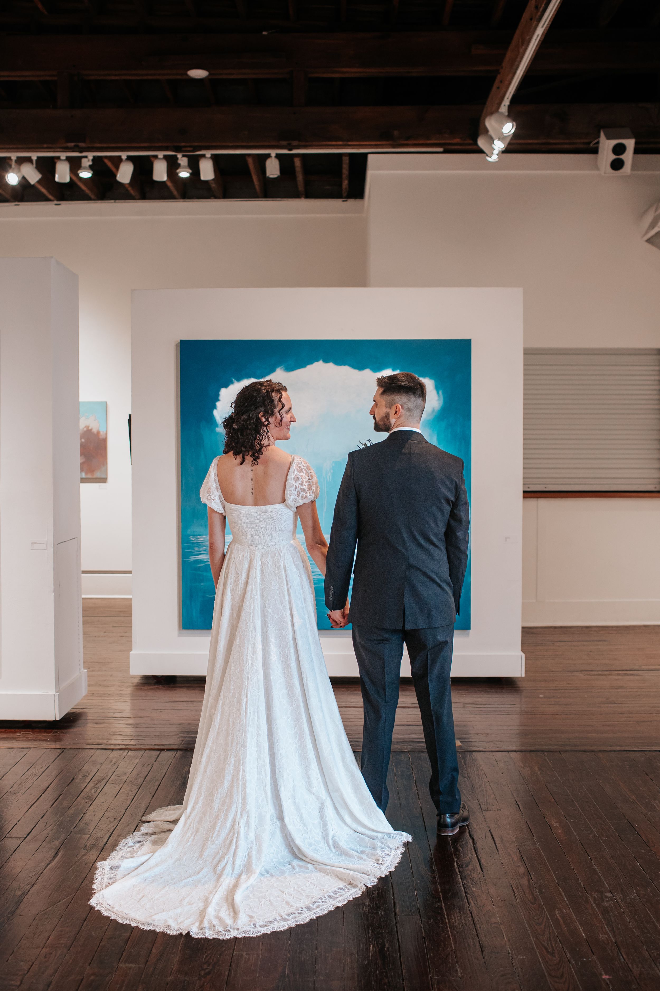 A couple in wedding attire stands together viewing artwork in a modern gallery with hardwood floors and track lighting.
