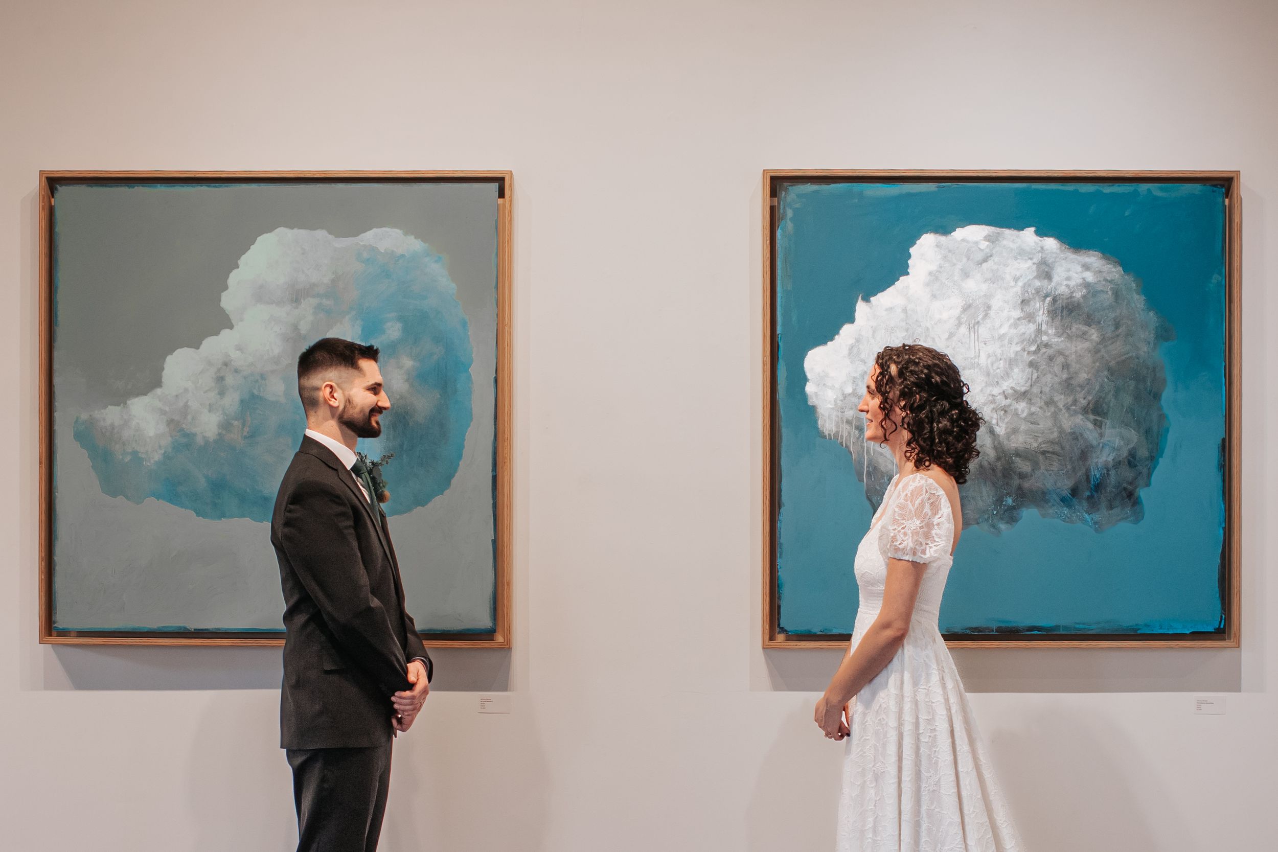 Two people in wedding attire stand facing cloud paintings on a gallery wall with blue and gray backgrounds.