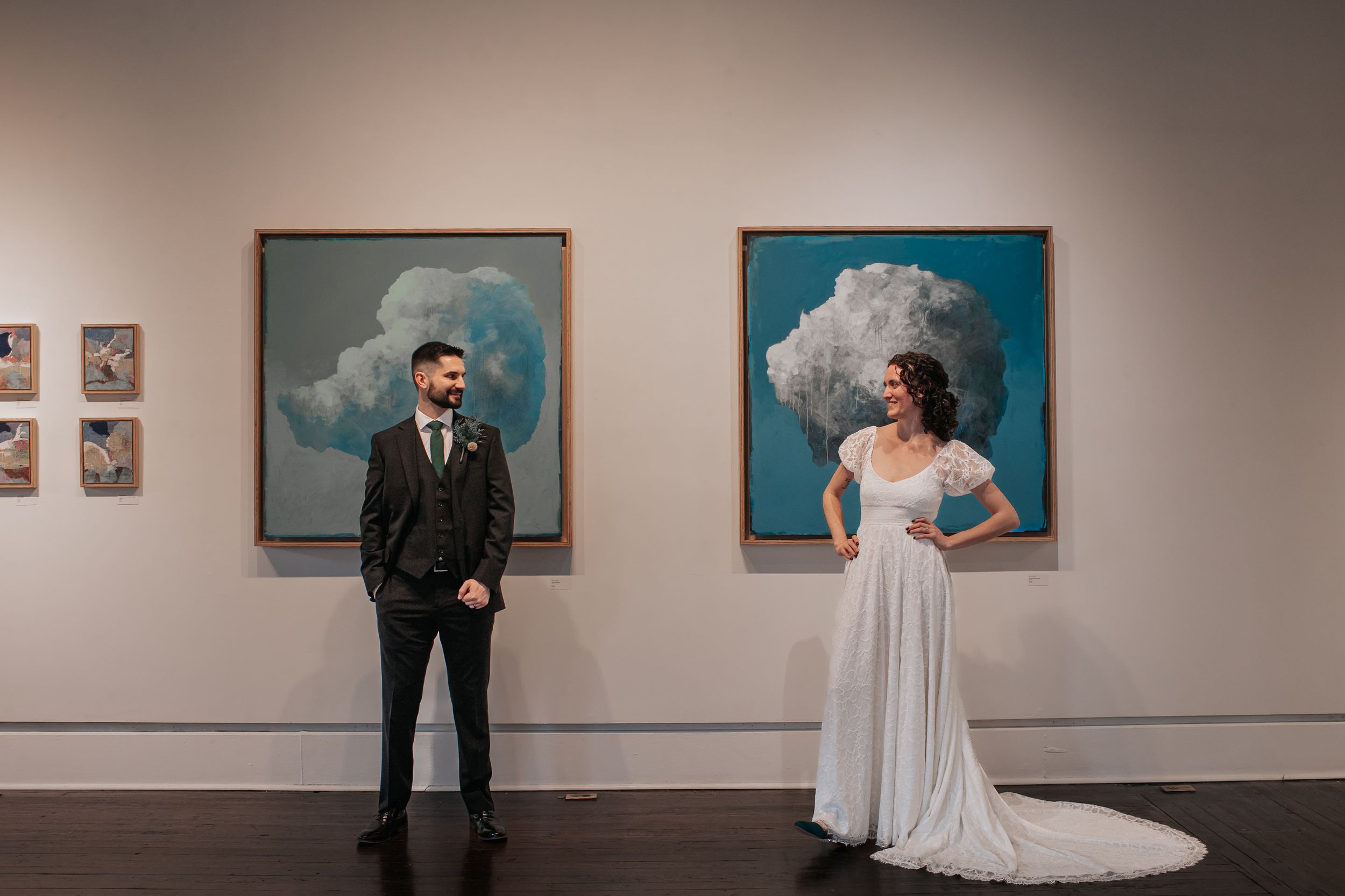 A couple in formal wedding attire poses in an art gallery with framed artwork displayed on white walls.