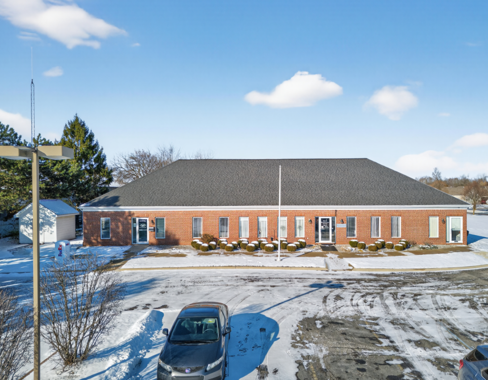 Brick single-story commercial building with dark roof surrounded by snow-covered parking lot on a sunny winter day.