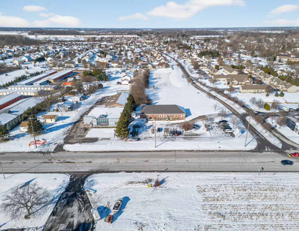 Aerial view of a snow-covered suburban area along a main road with a brick building and residential neighborhood.