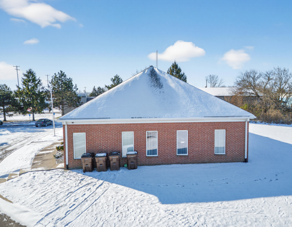 Snow-covered brick building with hip roof, multiple windows, and trash bins outside.