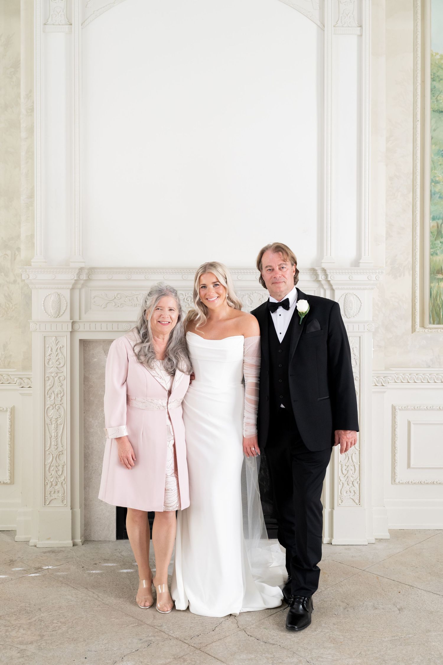 A wedding party poses against a white wall in an elegant room with ornate architectural details and natural lighting.