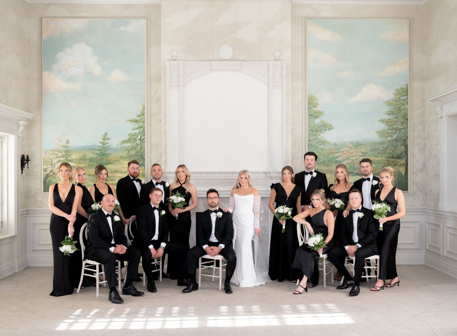 A wedding party in black and white formal attire poses together in an elegant room with painted landscape murals on the walls.
