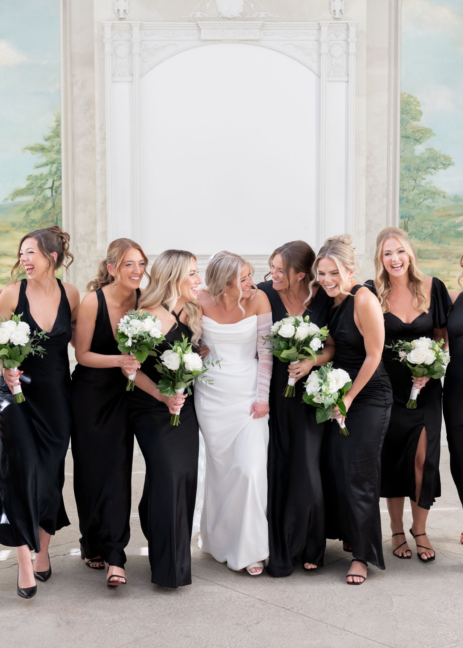 Wedding party in black dresses and white gowns pose together while laughing and holding white floral bouquets.