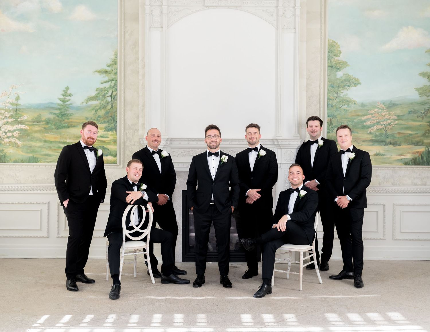 A formal group in black tuxedos poses together in an elegant room with a painted landscape backdrop.