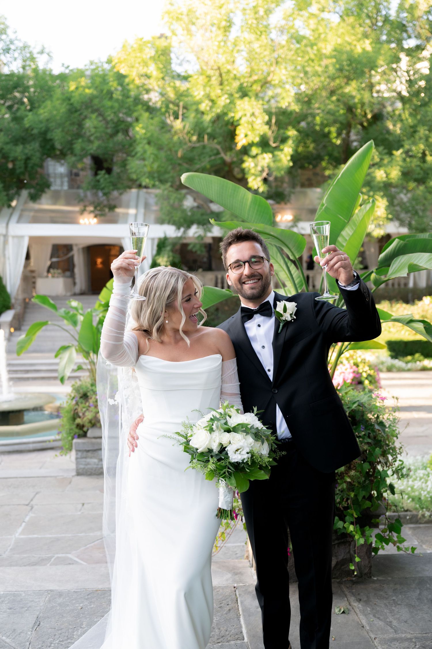 A couple celebrates joyfully in formal wedding attire in a garden setting with greenery and stone pathways.