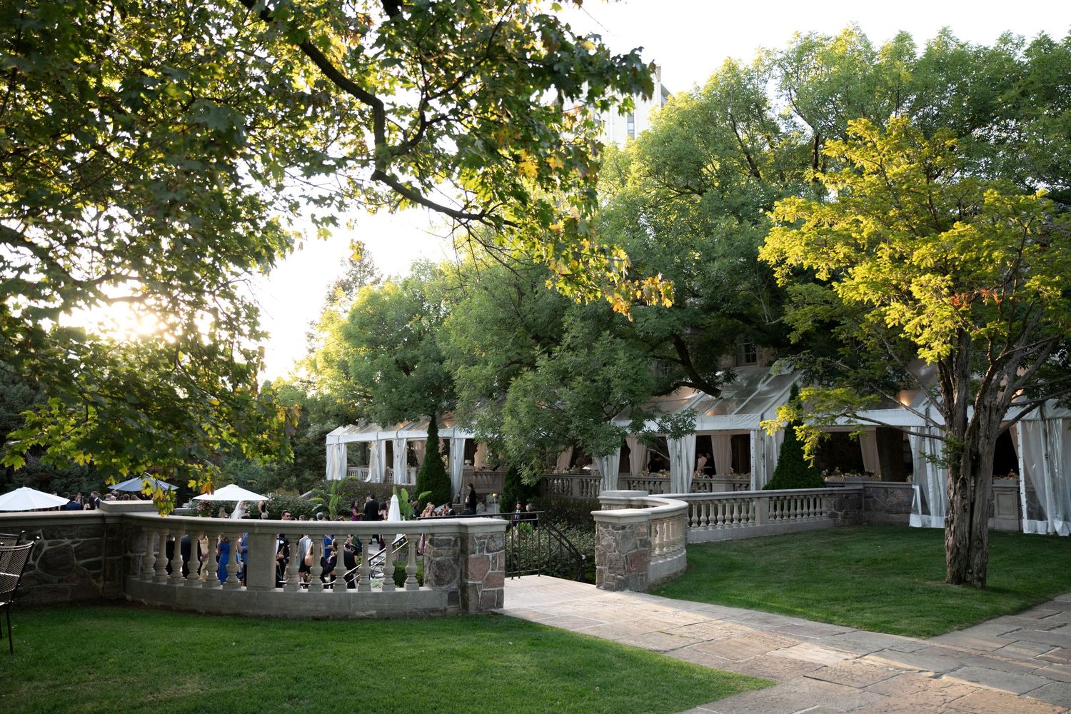 Elegant garden terrace with stone balustrade and white buildings surrounded by lush trees at sunset.
