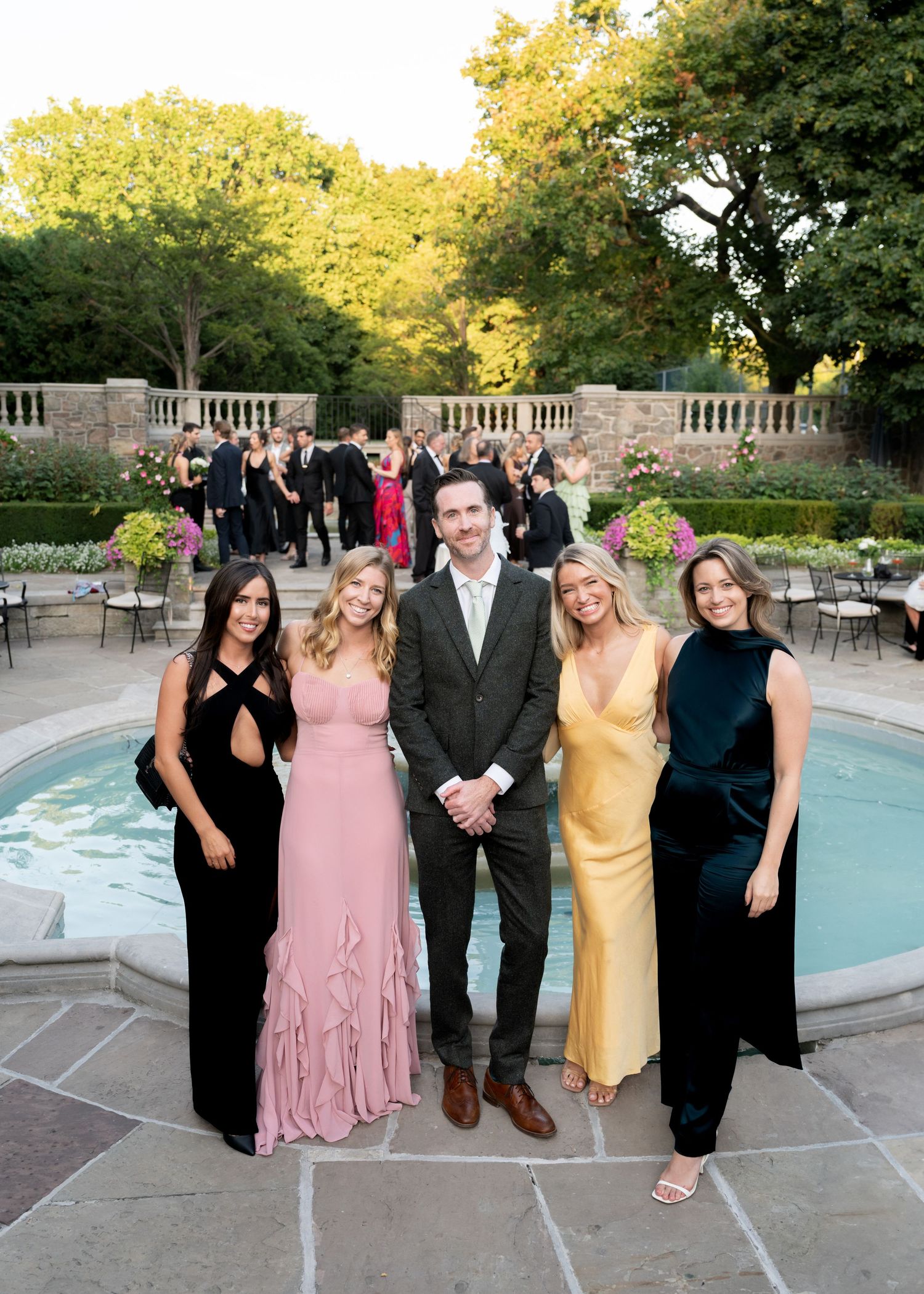 Group of friends in formal attire pose together at an outdoor garden party by a fountain at sunset.