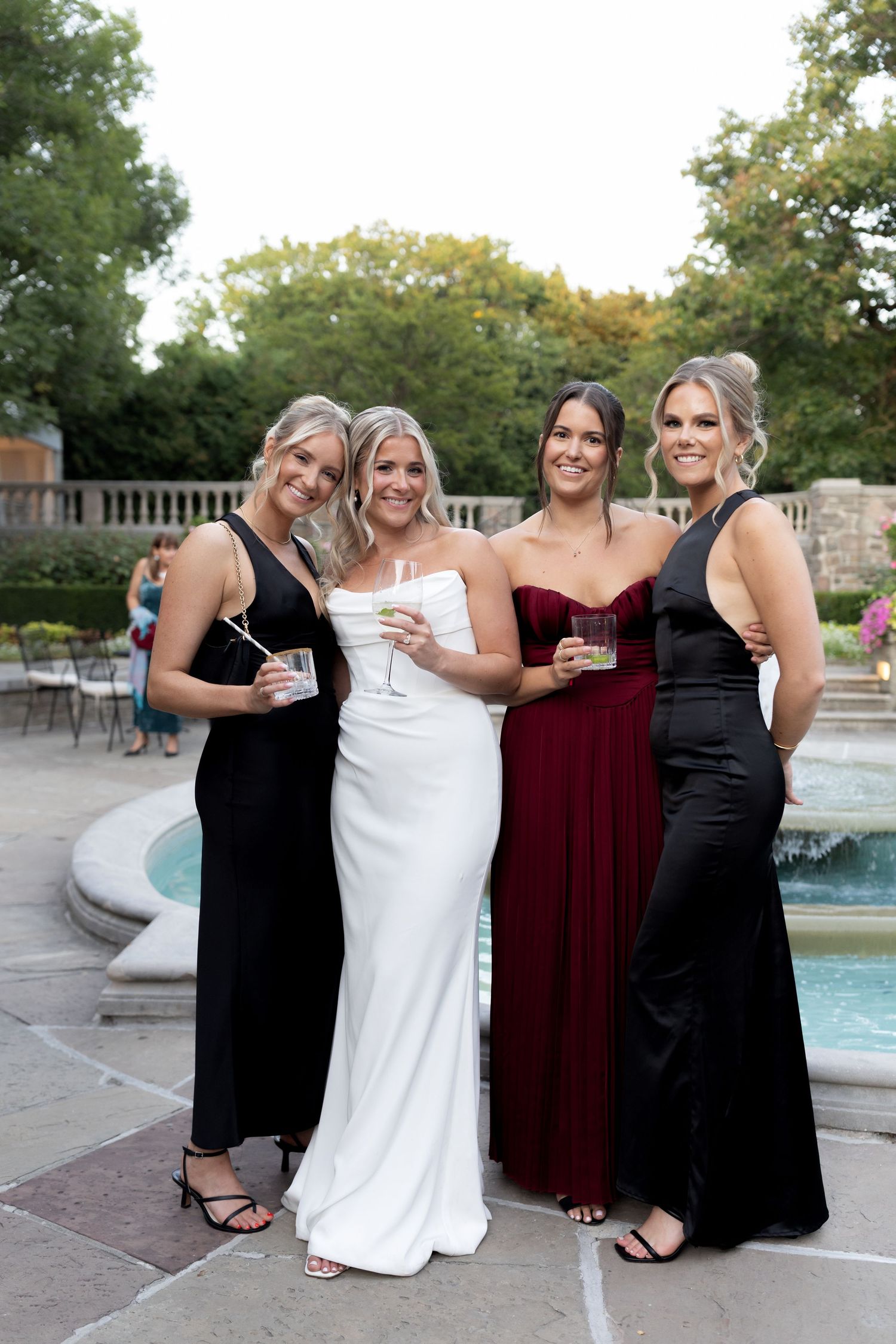 Four friends in formal attire share laughs and smiles while posing together by a pool during an outdoor celebration.