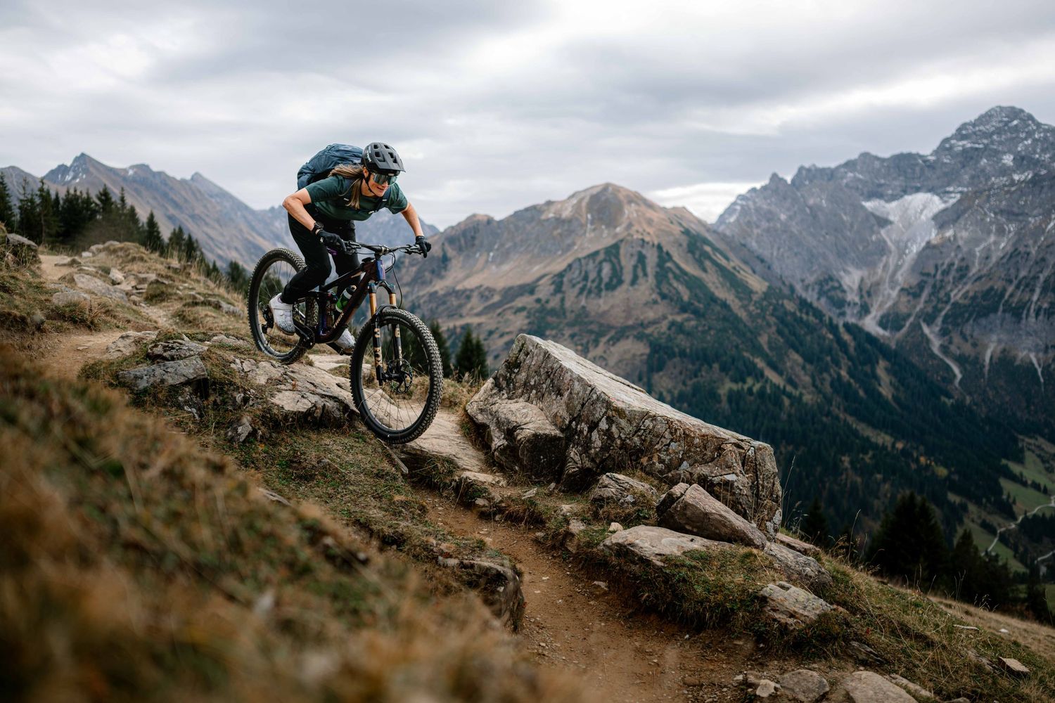 Mountain biker navigates rocky alpine trail with dramatic mountain peaks and cloudy skies in the background.