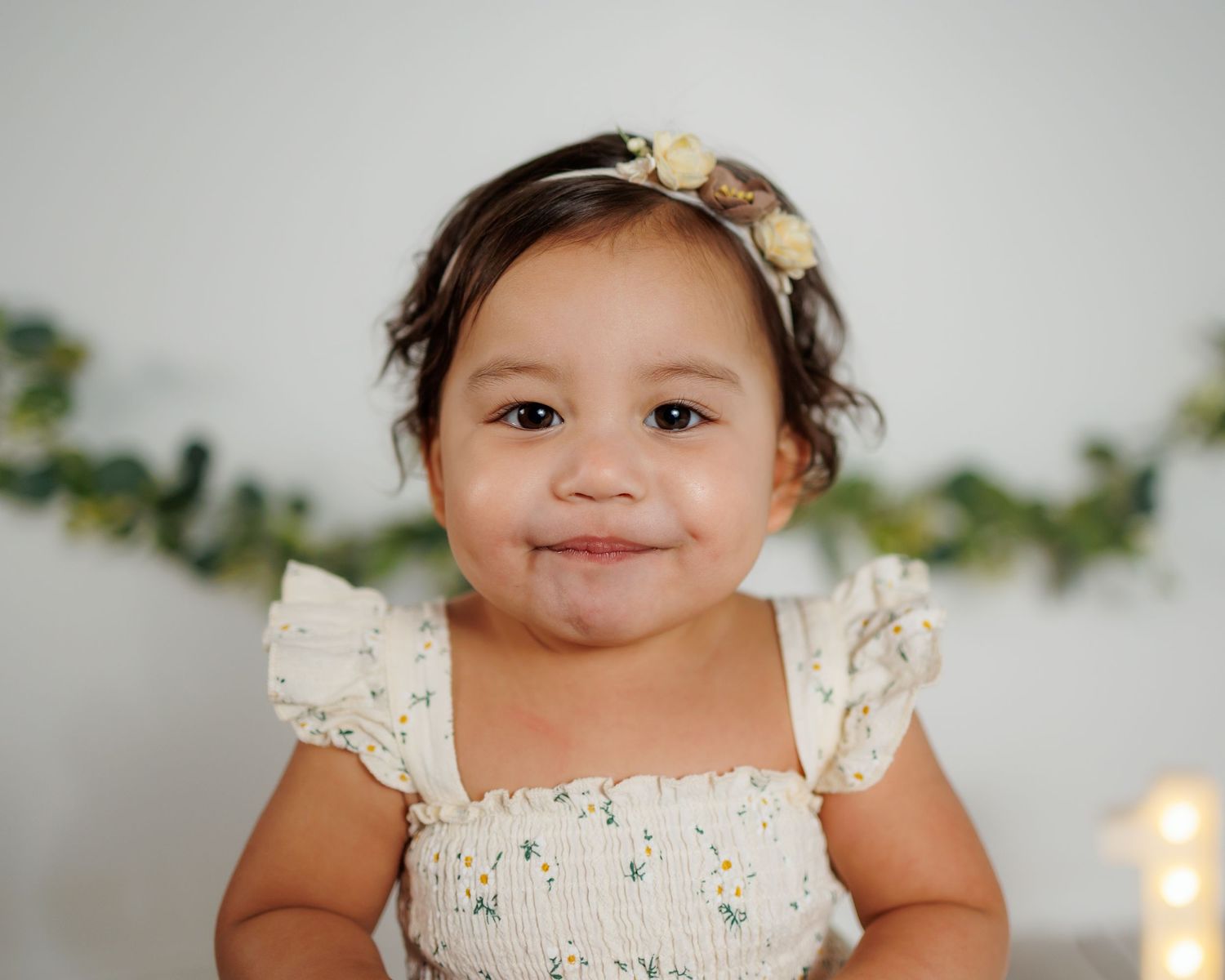 A baby wearing a cream-colored lace dress smiles sweetly for her first birthday photoshoot in a cozy studio in Attica, Michgan.