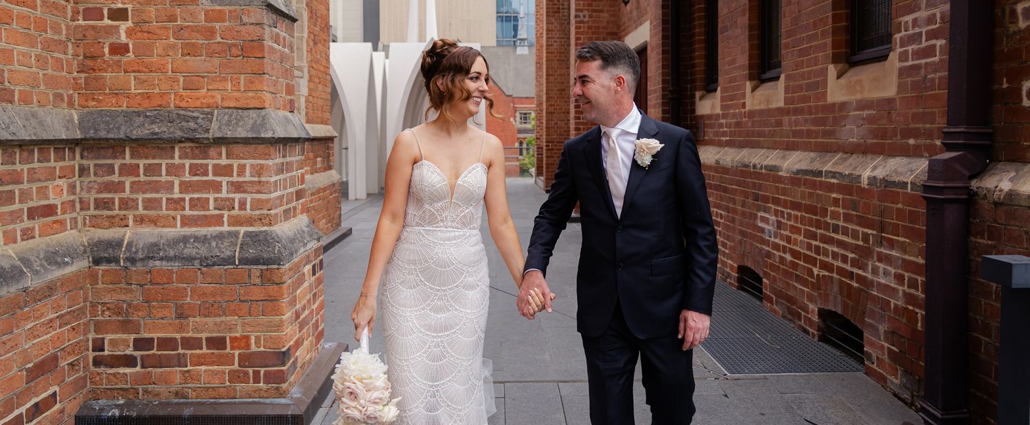 A couple in formal wedding attire walks along a grand building exterior with classic columns and architecture.