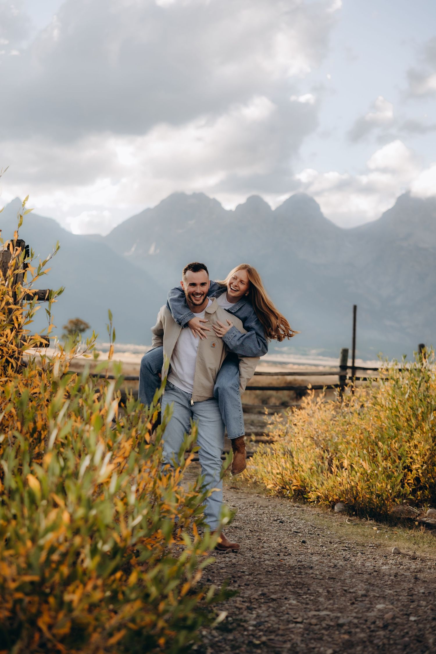 A couple embraces on a mountain trail with dramatic mountain peaks and cloudy skies in the background.